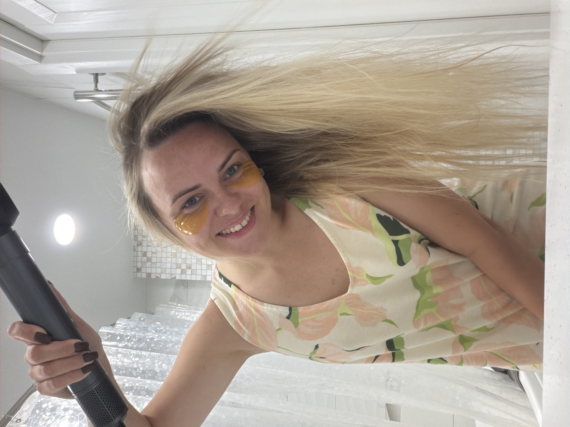A woman with blonde hair, wearing eye patches and a floral-print dress, is holding a hairdryer to her hair. She is smiling and looking at the camera. The scene appears to be a bathroom, with a shower curtain and tiled walls visible. The image was taken in Votkinsk, Russia.