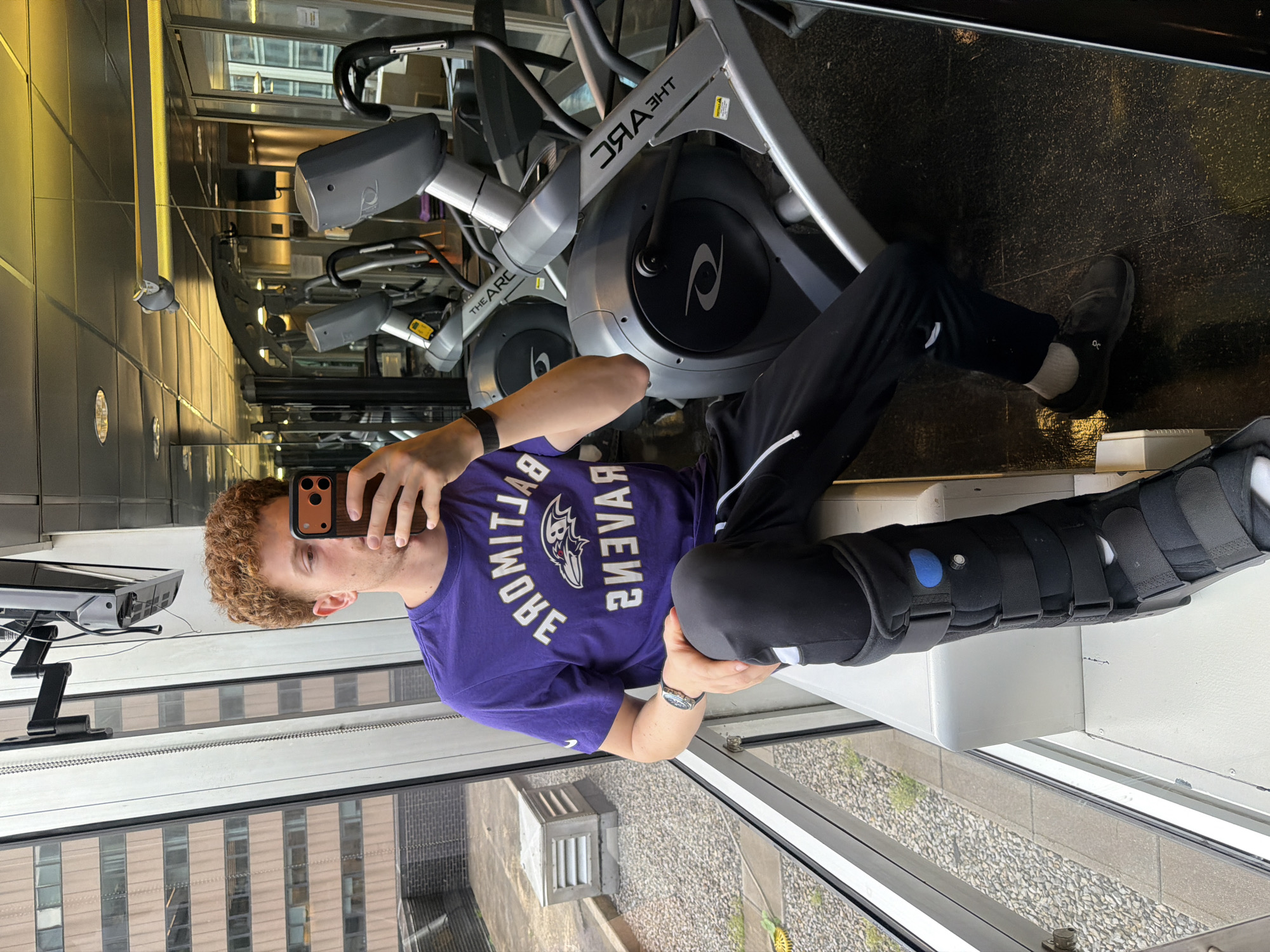 A fair-skinned person with curly red hair, wearing a purple Baltimore Ravens T-shirt, black sweatpants, and a black walking boot on their left leg, is taking a selfie with a phone in a room that appears to be a gym. The person is seated on a white surface near a large window, with elliptical machines behind them. Outside the window, there is a view of a city, possibly New York City, based on the building architecture. The person has a watch on their left wrist and a black band on their right wrist. A wall-mounted TV is visible to the left of the person. The floor is a dark color, and the lighting in the room is bright.