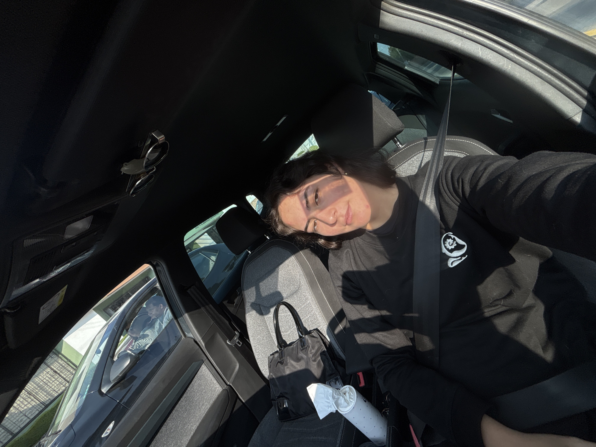 A young woman with short, dark hair is captured in a selfie or portrait taken from inside a car. She is seated in the front passenger seat, wearing a black long-sleeved shirt with a white circular logo featuring an abstract design on the left chest. A seatbelt is fastened across her chest. Her face is illuminated by strong, direct sunlight entering through the window, casting shadows across her features, and she has a soft, slightly smiling expression.
The car's interior is visible, featuring dark-colored upholstery with a light grey textured pattern on the passenger seat. A black handbag/tote bag rests on the seat beside her, along with a white travel mug or cup containing some tissue or paper. Through the car windows, glimpses of the outside environment show other vehicles, a building, and possibly a fence, suggesting an urban or suburban setting. The bright sunlight indicates it is daytime and likely sunny. No explicit text is clearly visible on objects, only the graphic logo on her shirt. The scene captures a casual moment, possibly while traveling in Puebla, Mexico, as inferred from the provided context.