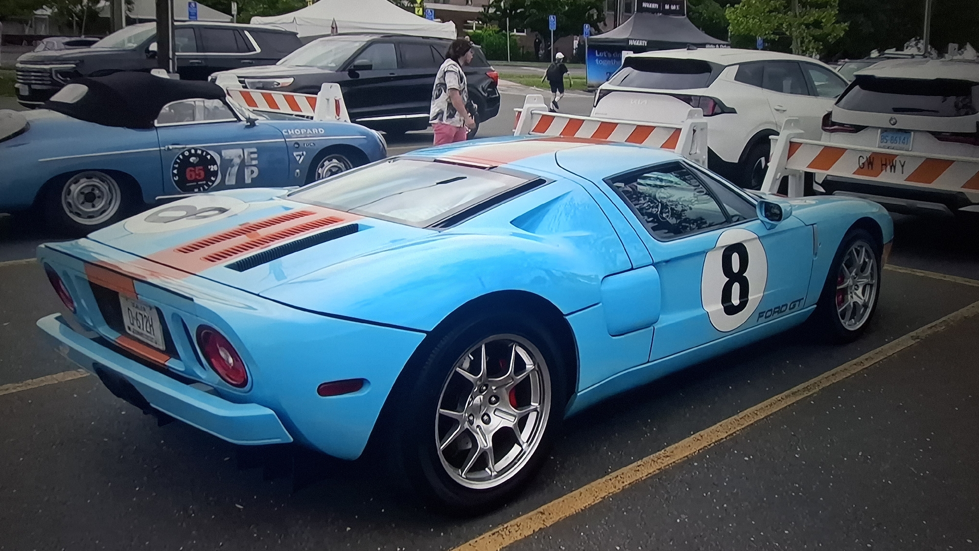 The image captures an outdoor car show or gathering, centered on a striking light blue Ford GT. This sports car features orange racing stripes across its hood and rear, with a prominent white circle containing the number "8" on its side. Its rear license plate reads "D-672H".
Behind the Ford GT, a light blue convertible classic car, possibly a Porsche 356 Speedster, is visible. It is adorned with various racing decals, including "CHOPARD", "CALIFORNIA 75", "65", "EP", "TOCS", and "365". In the background, several other vehicles are parked, including dark and white SUVs. A white BMW SUV displays the license plate "BS-66141".
The setting is a paved parking lot with yellow lines, likely at an event, as indicated by large white tents, one of which partially shows text like "HAGGARTY" and "Let's do together". Orange and white striped barrier fences separate some vehicles; one barrier is marked "GW HWY". People are seen walking in the background, including a man in a patterned shirt and pink shorts, and another individual further away. The event takes place during daytime, under what appears to be overcast or diffused lighting conditions.