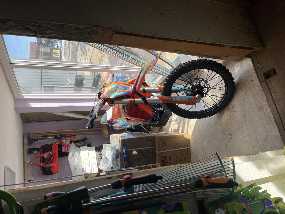 The scene depicts the interior of a structure, identified as being in Popovka, Russia, characterized by a mix of storage and display elements. A prominent orange and iridescent off-road motorcycle, consistent with an enduro or dirt bike, is positioned on a light-colored floor, angled towards a large glass door or window unit. The motorcycle features a black front wheel with a spoked rim and disc brake, orange front fender, forks, and frame components, alongside multi-colored, holographic-effect fairings.
To the left of the motorcycle, a vertical metal shelving unit displays various gardening tools, including rakes with black and orange handles and multiple green spray nozzles. Behind this unit, the back wall, painted a light lavender hue, features a wall-mounted grid panel from which a red fabric tool bag hangs. Several long-handled tools, including a broom, are leaned against the wall. Cardboard boxes, some explicitly labeled "DENZEL WATER PUMP," are stacked on the floor and against the left wall, alongside bundles of white, plastic-wrapped items.
Bright sunlight streams through the glass door/window on the right, casting distinct shadows on the floor and illuminating the motorcycle. Through the clear glass, an exterior environment is visible: patches of white snow or ice are present on the ground, indicating cold weather. Further back, a large building with gray corrugated metal siding and a brown-roofed structure are discernible under a clear blue sky. The immediate right wall within the interior appears unfinished or partially constructed, exposing insulation material. No individuals are present in the image.