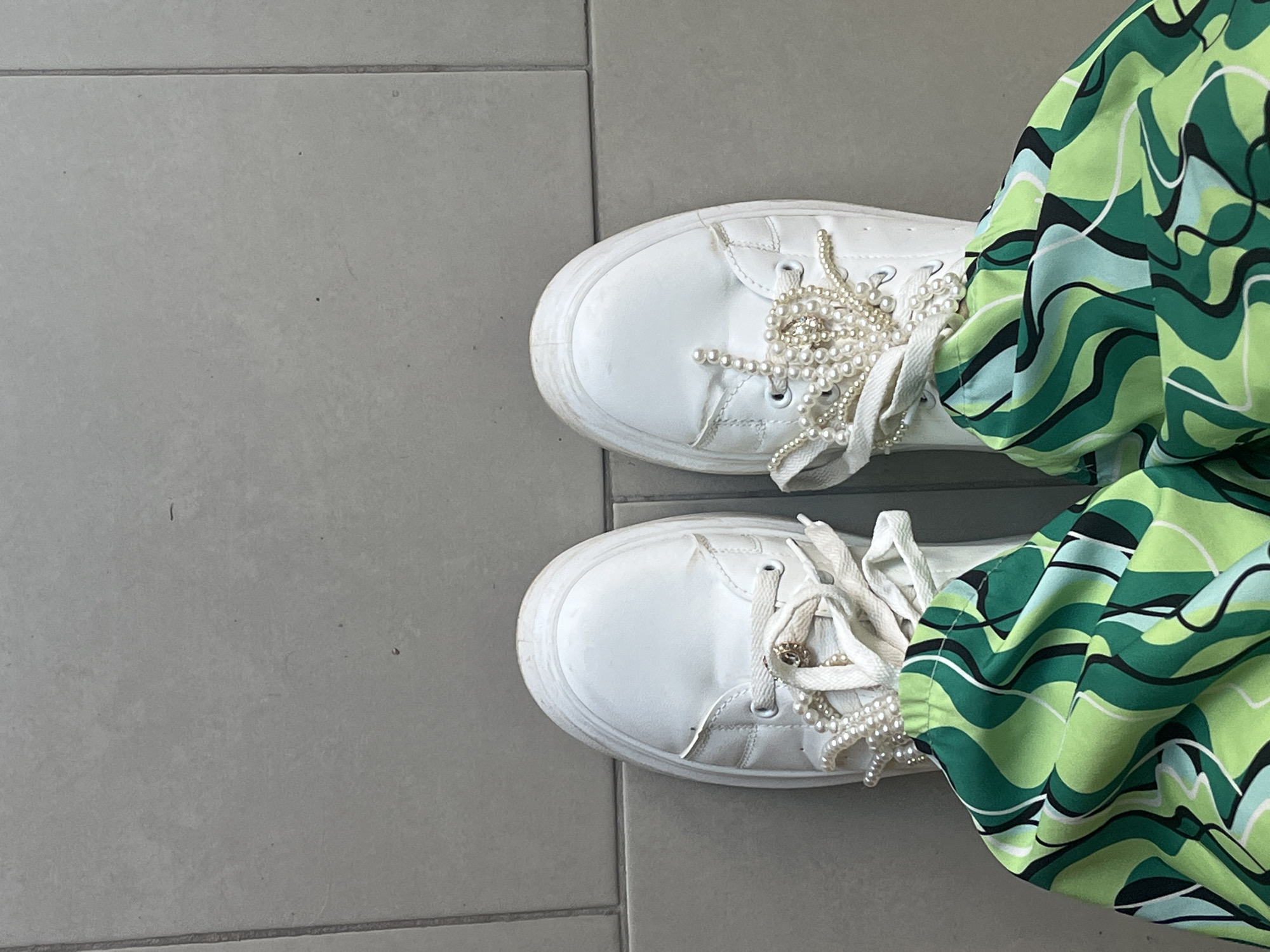 The image is a top-down shot of a person's feet standing on a gray tiled floor. The person is wearing white platform sneakers with pearl embellishments on the laces. The right side of the image shows a portion of patterned fabric, likely from pants or a skirt, featuring a wavy design in shades of green, black, and white. The location is Unknown, Unknown.