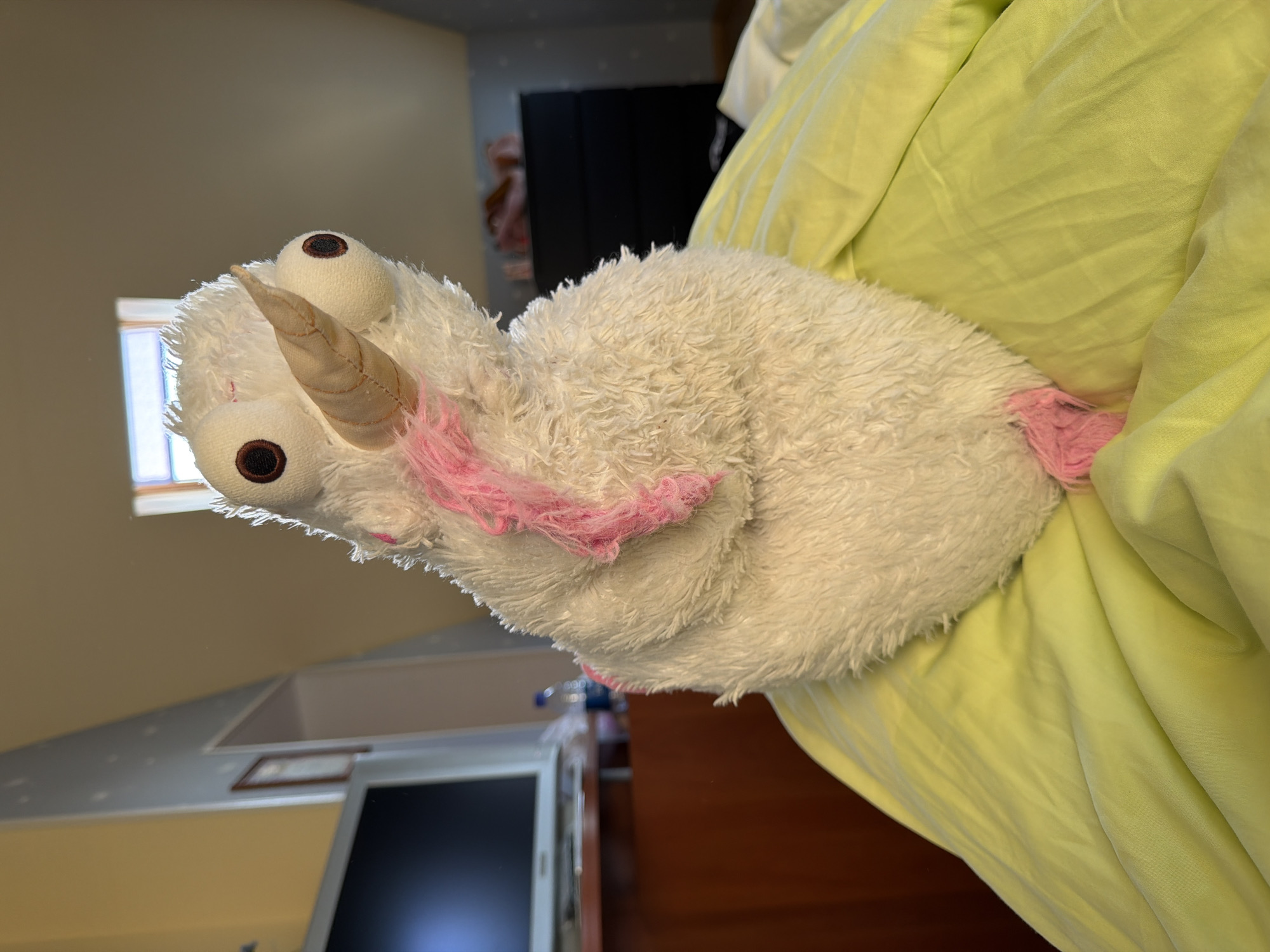 A close-up, eye-level, medium shot captures a white, plush toy with a conical horn and large, round, dark-brown eyes. Pink wisps of material, likely yarn, are attached to the toy's head and body. The toy is positioned on a lime-green surface that appears to be bedding. The background is softly blurred, showing a sloped ceiling, a window with a view of the sky, a dark dresser, and part of a wooden desk with a monitor on it. The lighting suggests an indoor environment, possibly a bedroom. The location of Жостово, Russia, is not visually discernible in the image.