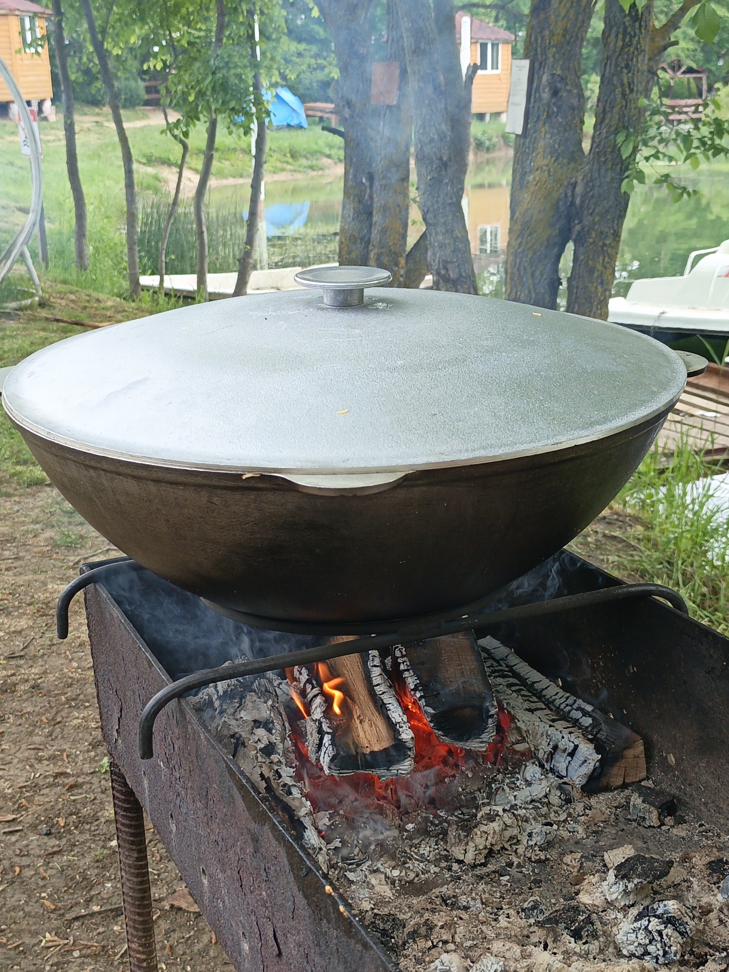 The image shows a large, dark-colored cast-iron pot with a light gray lid sitting atop a metal barbecue grill. The grill contains burning wood and hot coals, with smoke rising from the fire. The setting appears to be outdoors near a body of water. In the background, there are trees, a lake reflecting the sky and buildings, and some structures that look like cabins or houses. The location is Ternovskyi okrug, Ukraine.