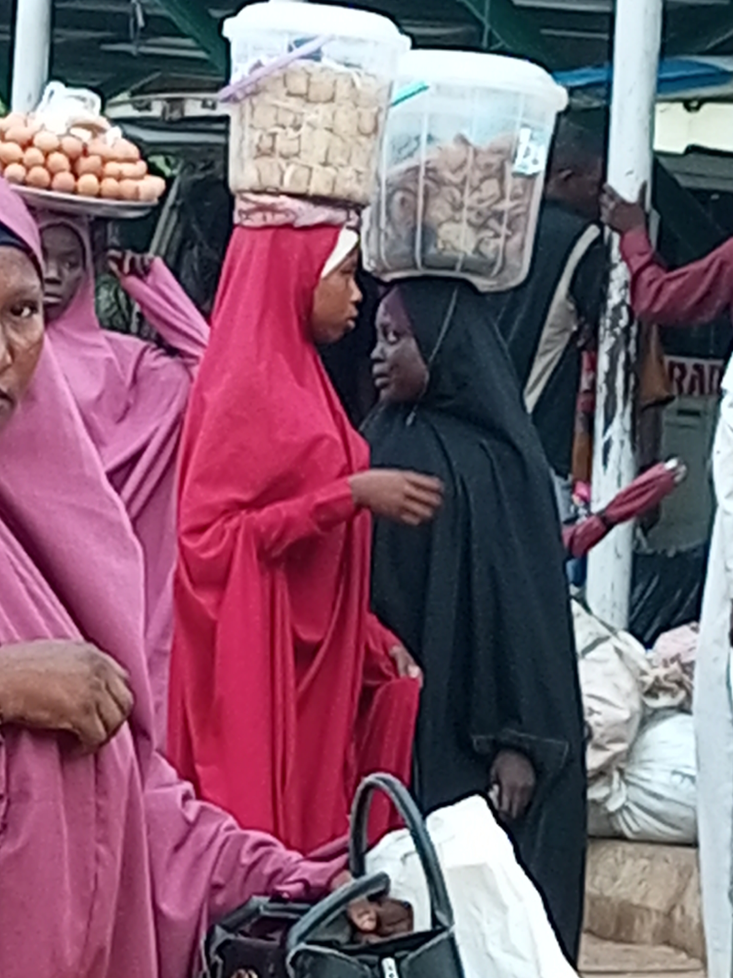 The image depicts a busy outdoor market scene in Gombe, Nigeria. In the foreground, a woman wearing a pink abaya is seen from the chest up, with her face partially visible. To her right, another woman, dressed in a vibrant red abaya, carries a large white plastic container filled with light brown cubes on her head. Her face is also partially visible. Behind them, a person in a black abaya and hijab stands, also with a plastic container on their head, this one filled with a lighter colored, granular material. To the left of the woman in pink, another person in a pink abaya is visible, also from the chest up. In the background, there are stacks of goods, including what appears to be eggs on a tray being carried by someone. The overall environment suggests a bustling marketplace with people engaged in commerce.