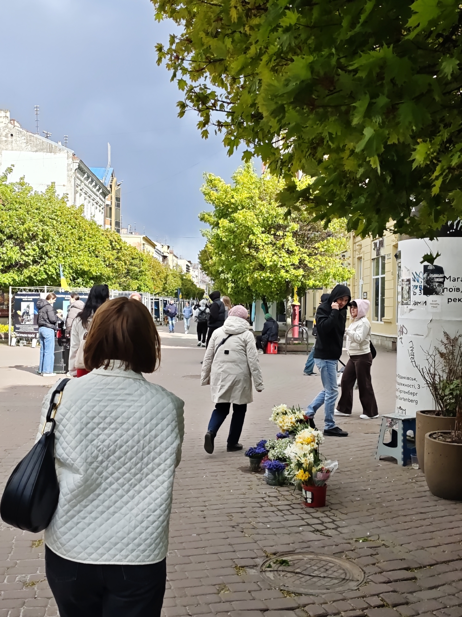 The image captures a street scene in Ivano-Frankivsk, Ukraine. The perspective is from behind a woman wearing a white quilted jacket and a black shoulder bag, who is facing down a brick-paved street lined with trees. A row of people are walking in both directions, some wearing light jackets and one in a dark hoodie. A small assortment of flowers is displayed on the ground for sale near a makeshift table and potted plants. Buildings stand along the street on both sides, and posters are affixed to a vertical structure to the right of the scene. The sky is overcast with clouds. The poster on the right advertises the "Magazyn" restaurant and includes contact details, specifying Ivano-Frankivsk, Independence Street, 3, Gartenberg.