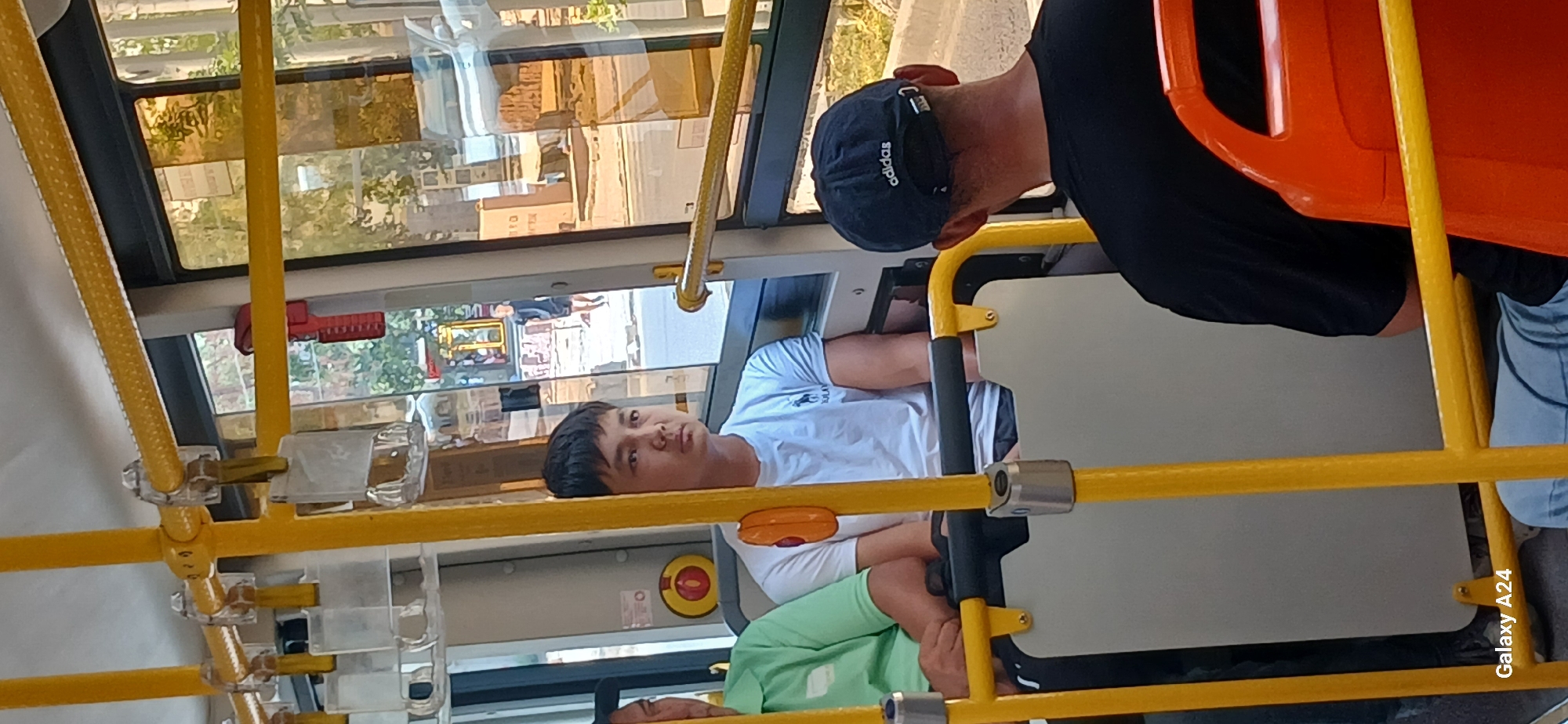The image depicts the interior of a public bus in Bishkek, Kyrgyzstan. Three individuals are visible. In the foreground on the right, a person wearing a black t-shirt and a navy blue Adidas cap is seated, facing away from the camera. To the left of this person, a young man in a white t-shirt with a dark logo is seated, looking forward. To the far left, another individual, only partially visible, is wearing a light green shirt and is seated. The bus features yellow metal poles and orange seating. The windows show reflections of trees and buildings, suggesting an urban environment outside. The "Galaxy A24" watermark is visible in the lower left corner, indicating the device used to capture the image.