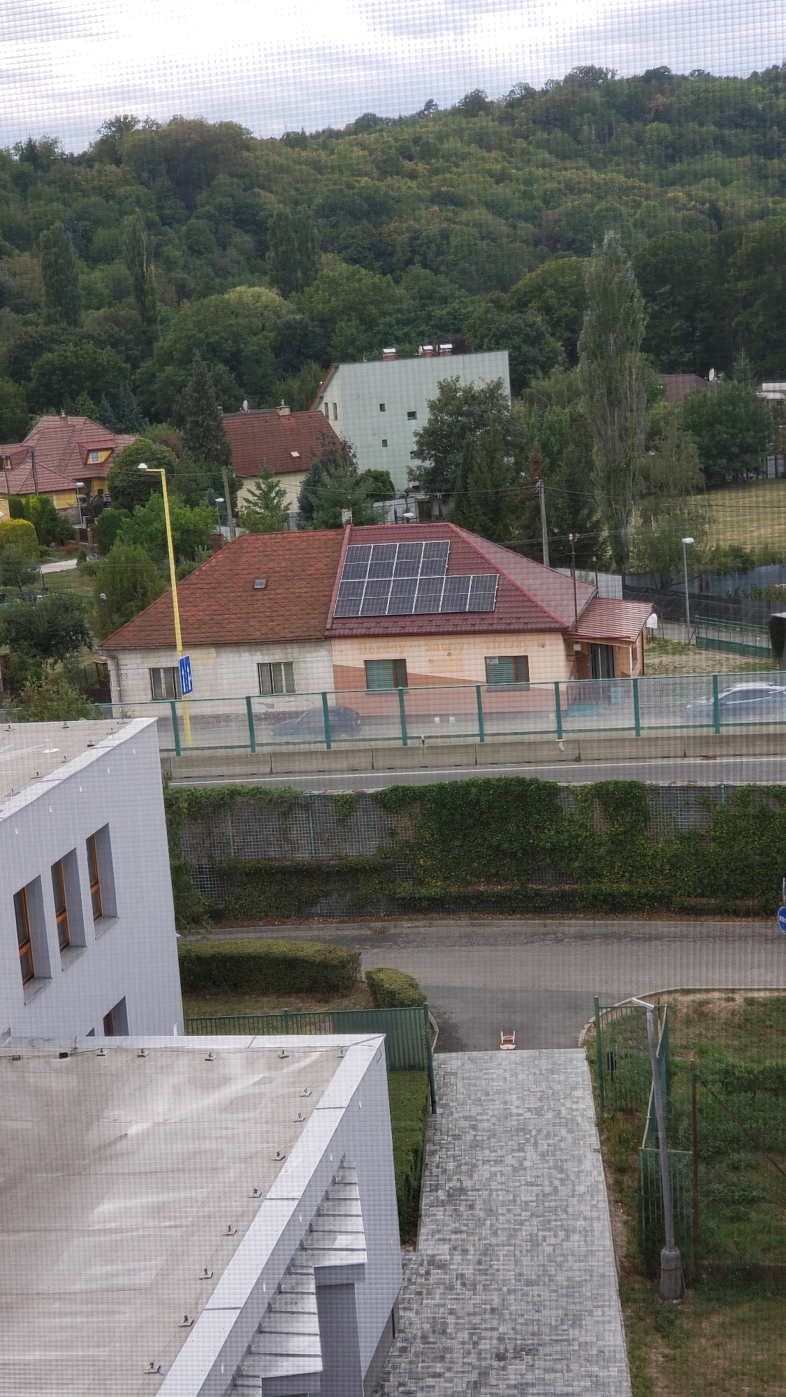The image captures an aerial view of a residential and wooded area. In the foreground, the roof of a modern building with light gray siding and a sloped section is visible. Adjacent to this is a paved pathway made of interlocking gray bricks. Further into the scene, a two-story house with a red tile roof is prominent, featuring a section of solar panels installed on the roof. The house has beige and white siding and several windows. Across from the house, a light green safety barrier runs parallel to a road. Beyond the road, a densely wooded hillside dominates the background, with a clear sky overhead. A streetlamp with a yellow pole stands near the house. The overall environment suggests a suburban or semi-rural setting. The image appears to be taken from an elevated position, possibly a window or a drone, looking downwards and slightly towards the east. The specific location within Prešov, Slovakia cannot be determined from the visual information alone.