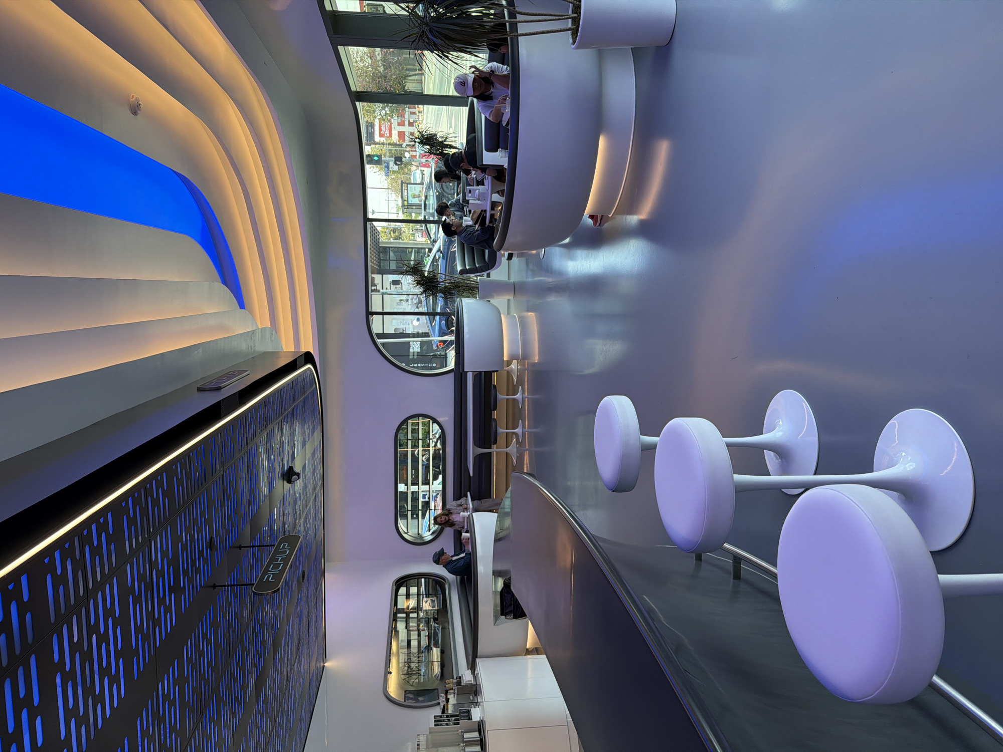The image depicts the interior of a modern cafe, likely located in West Hollywood, United States of America, characterized by its sleek, futuristic design. The space features a high ceiling with an intricate, perforated panel illuminated by blue light, contrasting with a wavy ceiling section bathed in warm, amber tones, accented by a vibrant blue strip. A prominent dark, curved counter dominates the foreground, with several white, tulip-shaped bar stools around it.
In the mid-ground, diners are seated in curved booths along a wall with distinctive oval windows offering views of the street outside, where cars and a traffic light are visible. A sign hanging from the ceiling indicates "PICKUP". There are multiple individuals, some appearing to be customers and others possibly staff, engaged in activities such as eating, drinking, and conversing. The floor is a highly reflective, light-colored surface that mirrors the ambient lighting. Potted plants add a touch of greenery to the otherwise minimalist environment. The overall impression is one of a clean, well-lit, and contemporary dining establishment.