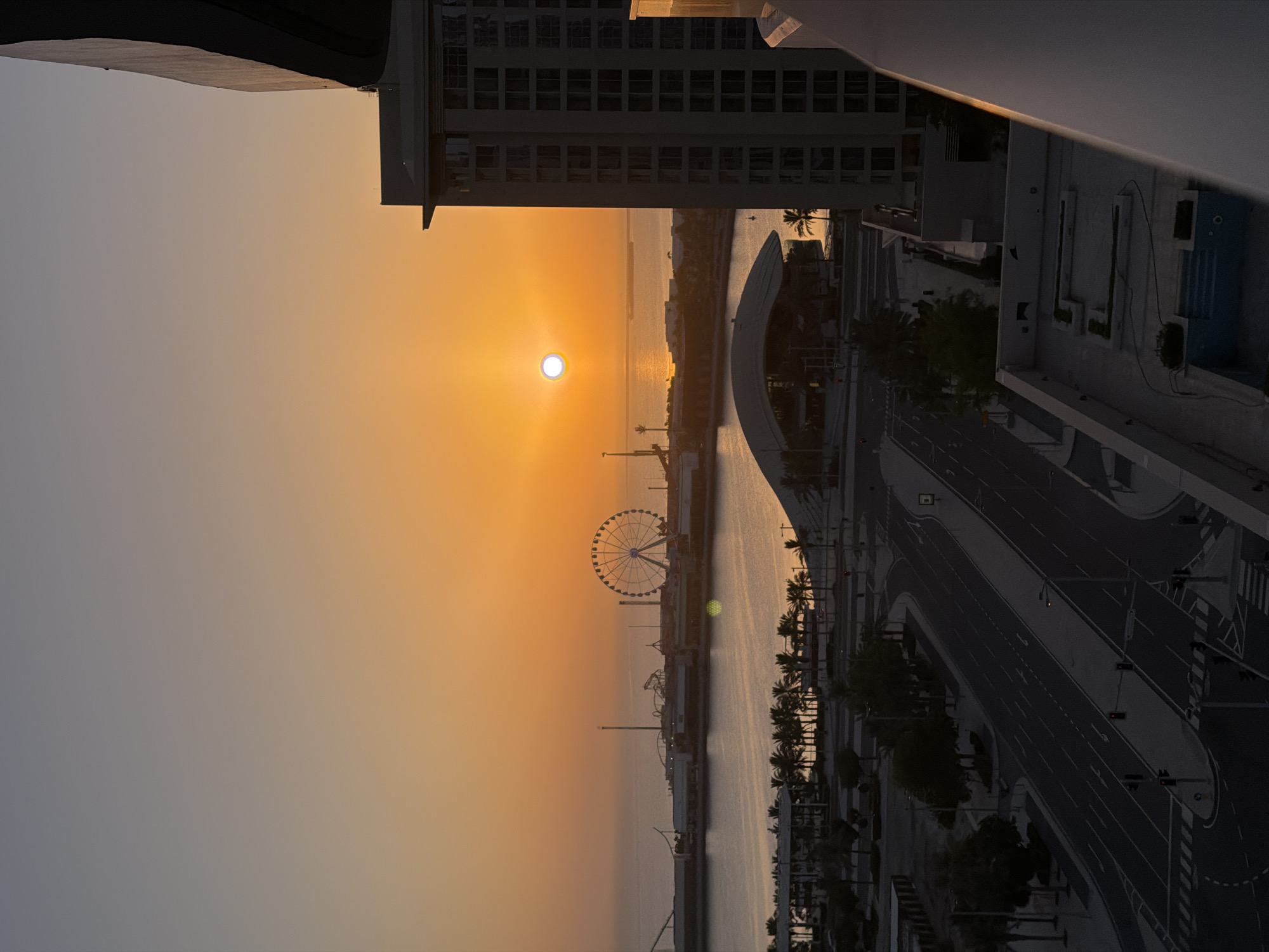 This image captures a sunset over a modern cityscape situated near a body of water. The sun, a bright orb, is positioned on the left side of the frame, casting a warm orange and yellow glow across the sky. In the middle ground, a Ferris wheel stands prominently, its spokes visible against the colorful horizon. The scene includes a waterfront with a curved, modern architectural structure and a road with streetlights and crosswalks. Palm trees are scattered throughout the landscape, suggesting a warm climate. The buildings in the background are contemporary in design, with multiple floors and windows. The overall mood of the image is serene, evoked by the tranquil water and the soft light of the setting sun. The time of day is clearly sunset. There is no visible text in the image.