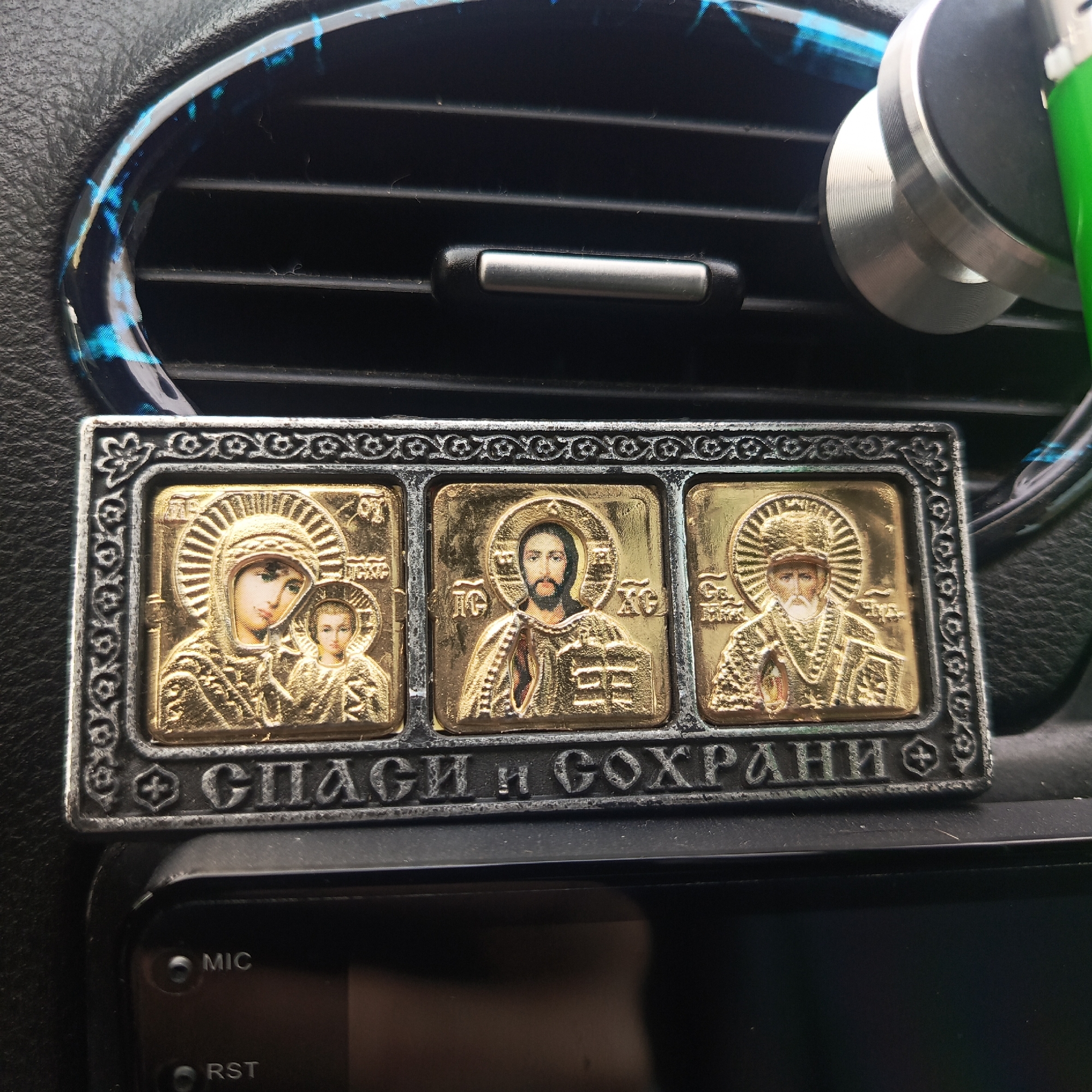 The image shows a religious icon in a car interior, specifically resting on the dashboard beneath an air vent. The icon is rectangular and features three panels depicting religious figures in gold relief against a dark metal background with a decorative border. The text "СПАСИ и СОХРАНИ" is inscribed below the panels. A smartphone car mount is visible to the right of the icon, attached to the air vent. The dashboard appears to be made of a dark, textured material. The location of the image is in a car, and since no information about the city of Chaikovsky, Russia is present, it is impossible to verify.