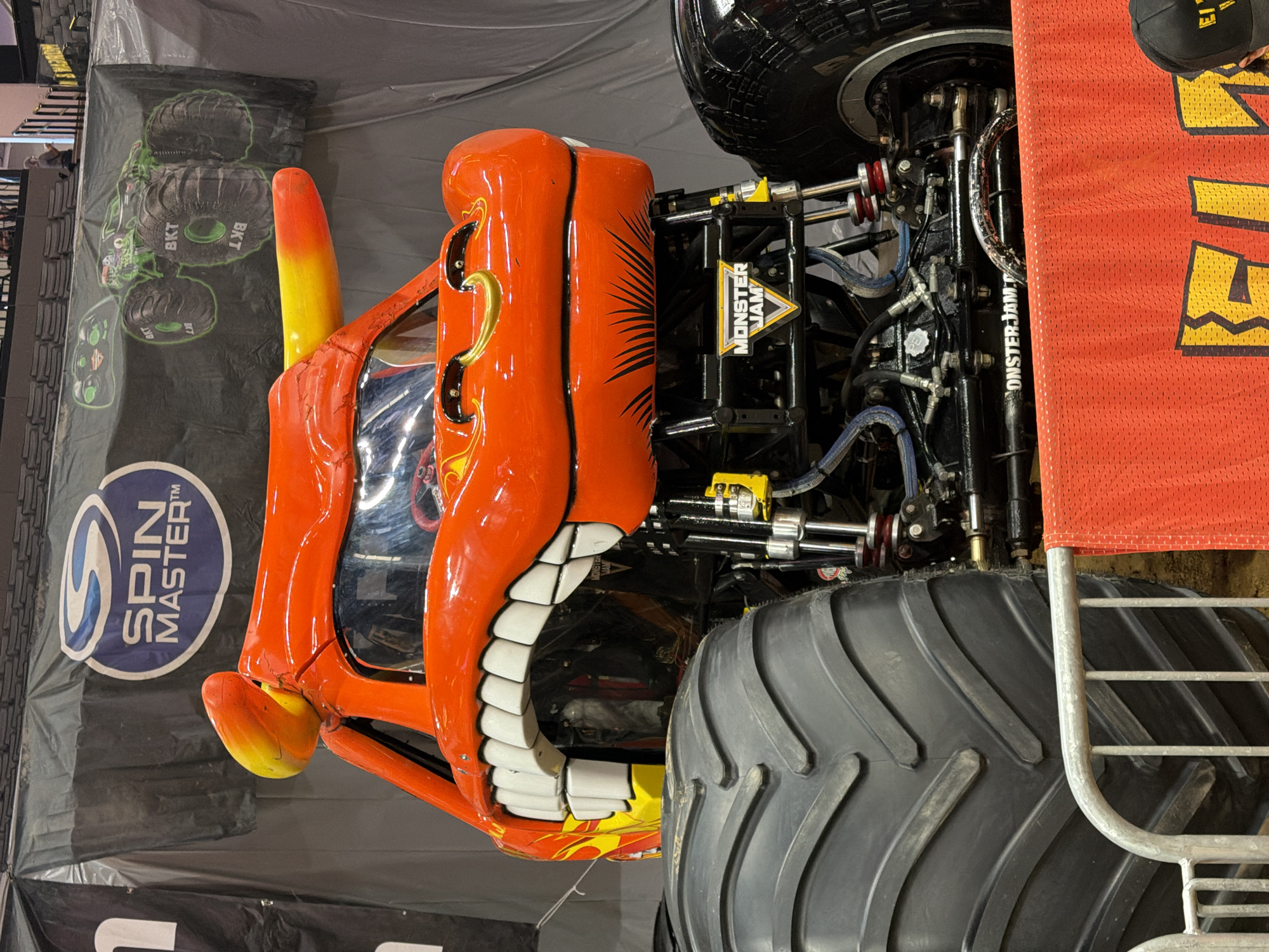 The image shows a close-up view of a monster truck adorned with a dragon-like face, painted in vibrant orange with yellow horns. Its large, white teeth are prominently displayed in a wide grin. The truck has massive black tires, with the front-left tire being the most visible, taking up a significant portion of the left side of the frame. The monster truck is parked in an indoor venue, likely an arena or exhibition hall, indicated by the presence of stadium seating in the background and a large banner hanging on the wall. The banner features logos for "Spin Master" and "Monster Jam," the latter also appearing on a smaller sign attached to the truck's chassis. The ground appears to be dirt or sand, typical for monster truck events. A metal barrier with an orange cloth draped over it is in the foreground, partially obscuring the truck's wheels and lower chassis. No people are clearly identifiable in the foreground, though a few figures can be seen in the distant background, seated in the stands. The scene is illuminated by artificial lighting. The location is identified as Sacramento, United States of America.