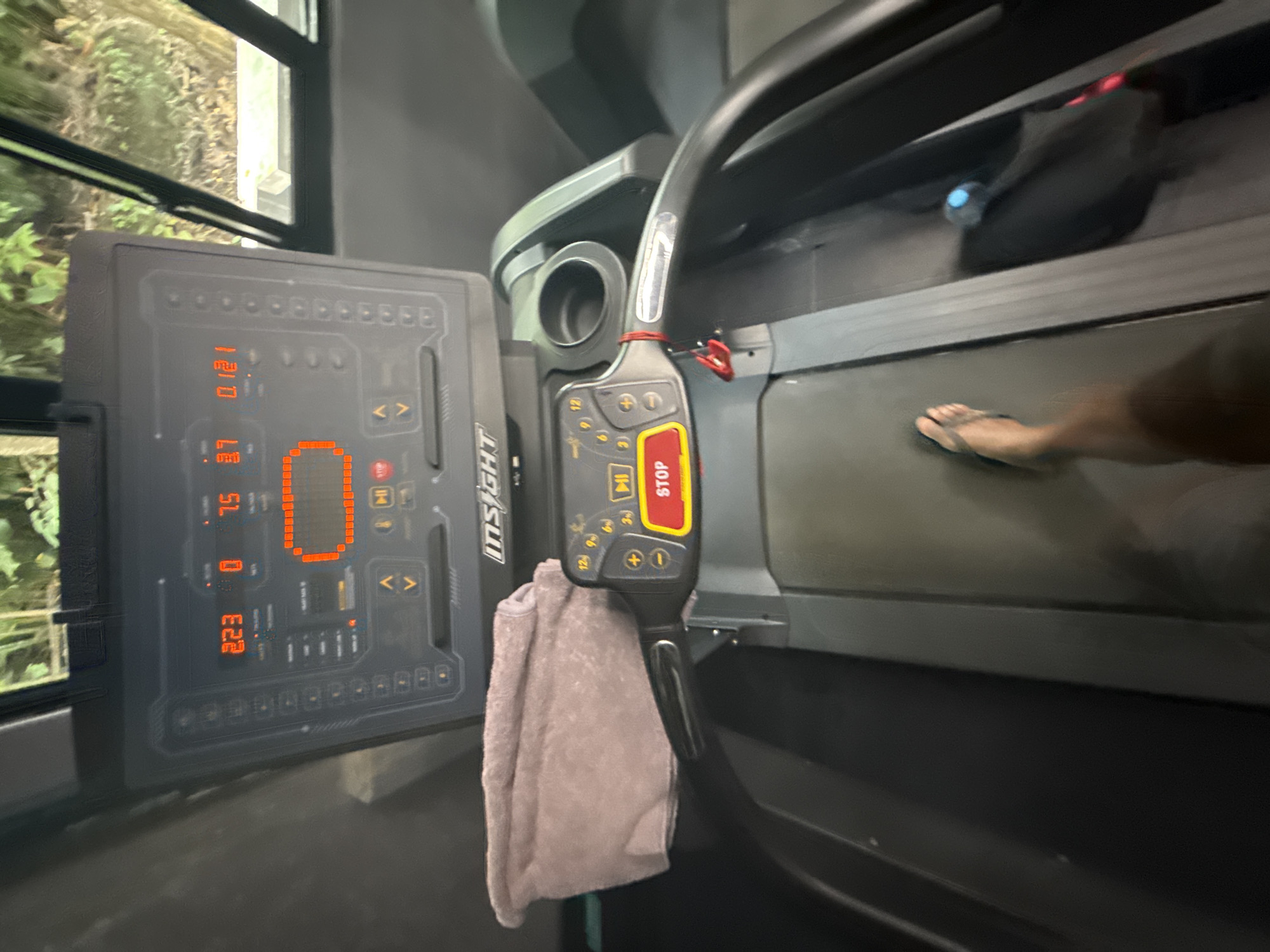 The image shows a treadmill with an "INSIGHT" brand display panel indicating 2.23 distance, 7.5 speed, and 12.1 incline. A towel hangs from the treadmill's handlebar, which features a prominent "STOP" button. A person wearing flip-flops is visible on the running surface. The location is Unknown, Unknown.