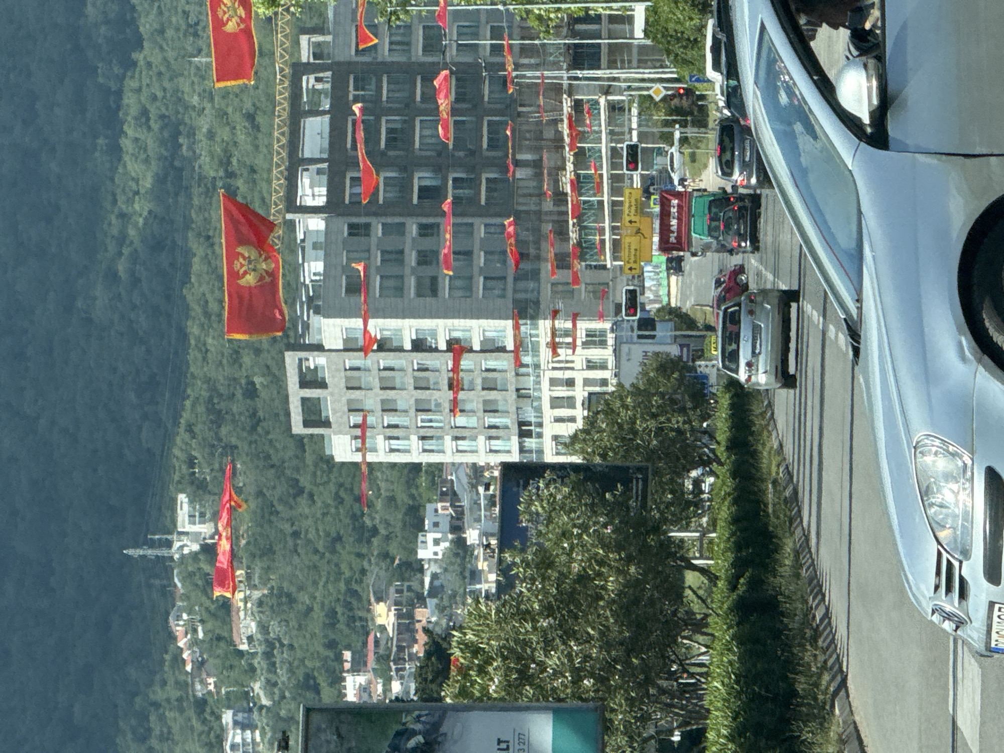 The image captures a street scene in Bar, Montenegro, lined with buildings decorated with red Montenegrin flags. A silver car occupies the right side of the frame, partially obstructing the view of the street. Behind the car, a line of vehicles is visible on the roadway, controlled by traffic lights. Buildings, adorned with numerous Montenegrin flags, flank the street. A wooded hillside rises in the background. The signage includes the word "PLANSEE," and directions to Podgorica, Rijeka and Cetinje.