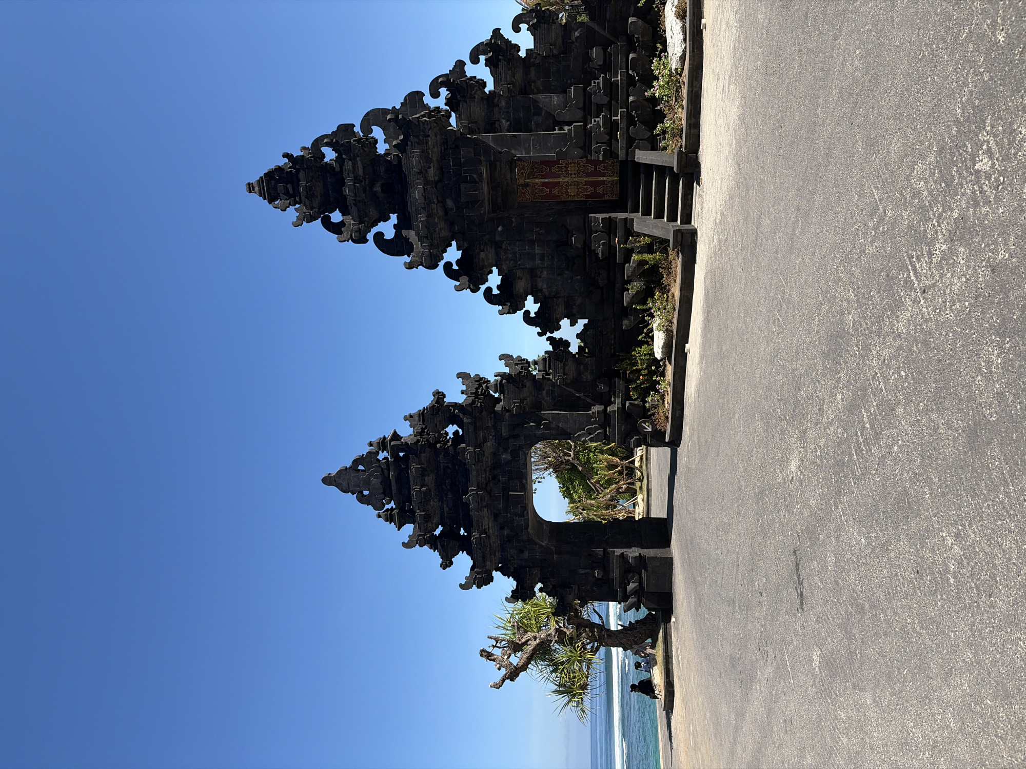 A wide-angle, eye-level, outdoor shot captures the iconic Pura Besakih's entrance gate, also known as the Lempuyang Temple or "Gateway to Heaven," located in Kuta Selatan, Indonesia. The imposing, dark stone gate, characteristic of Balinese architecture, stands proudly against a clear, vibrant blue sky. The gate is a split gateway, divided into two symmetrical structures, with intricate carvings and tiered roofs. A central archway allows a view of lush greenery and distant trees. To the left of the gate, near the ocean's edge, two individuals are seated on a low stone structure, observing the turquoise waters and gentle waves. A uniquely shaped tree with sparse foliage stands nearby, its branches reaching towards the sky. The foreground is an asphalt paved area, showing signs of wear and tear. The overall scene depicts a popular tourist destination, showcasing architectural grandeur and natural beauty.