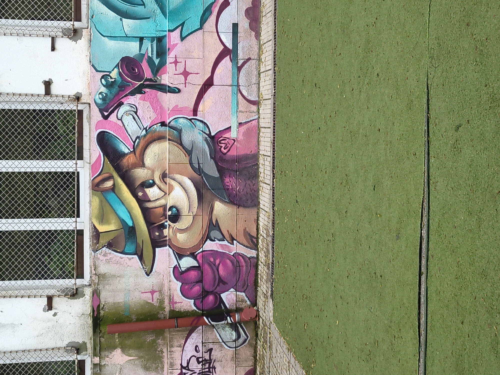 Captured from an aerial perspective, the image presents a combination of urban elements. The left side of the frame showcases a white building exterior, featuring a window with a wire mesh screen. Adjacent to the building, a vibrant mural adorns a wall, displaying cartoonish figures in hues of pink, brown, and turquoise. A rusty red pipe runs along the lower portion of the mural. To the right, a green surface, possibly a sports field or a similar recreational area, fills the remaining space, bisected by a narrow concrete seam. The scene appears to be located outdoors, potentially in a residential or mixed-use area within the city of Unknown, Unknown.