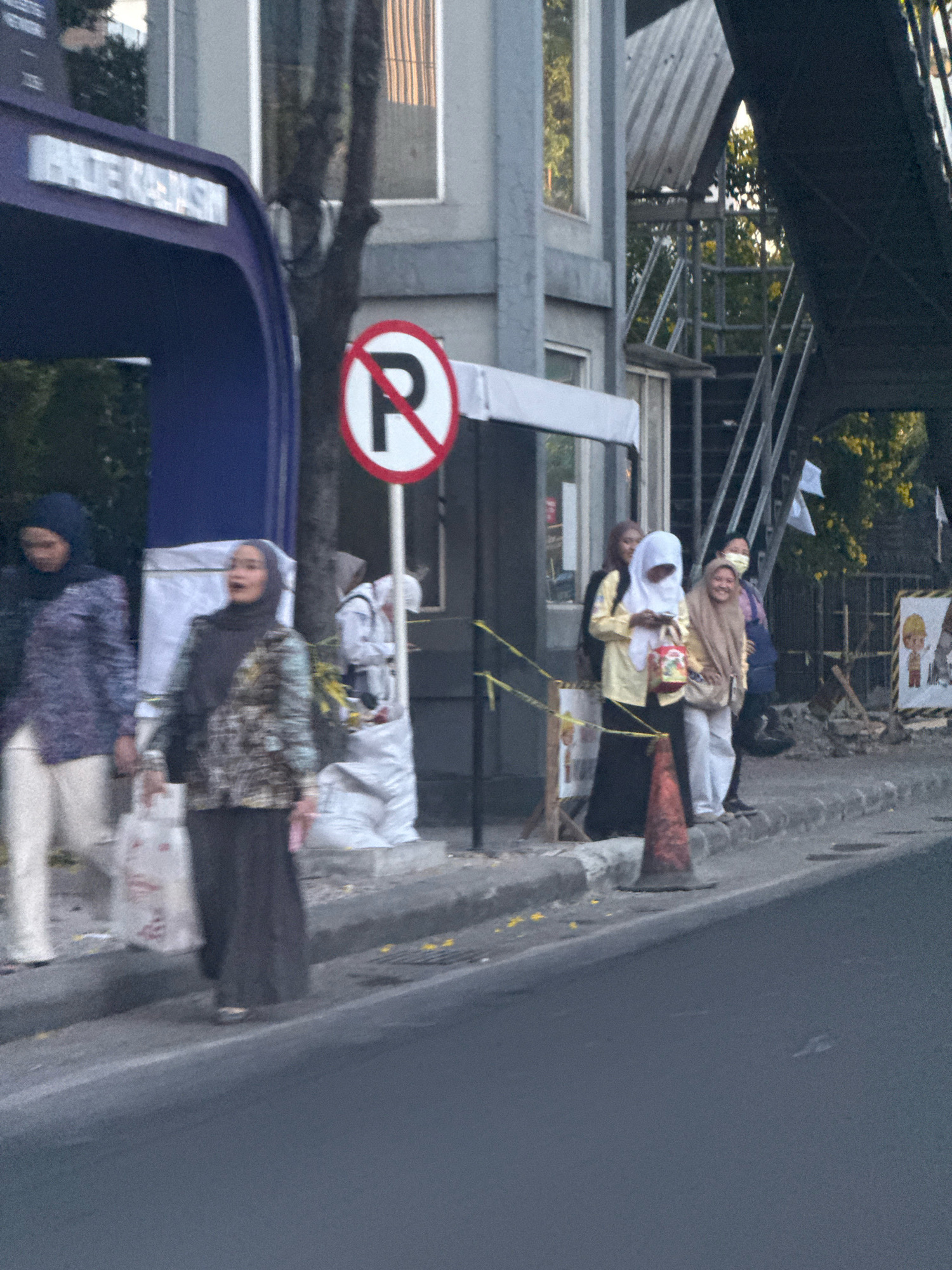 In Surabaya, Indonesia, a group of women are seen near a bus stop. One woman in the foreground, wearing a hijab and a patterned blouse, appears to be talking, with her mouth open. Another woman, also in a hijab, stands to her left, carrying a shopping bag. To the right of the first woman, a person in a white outfit is visible. Further to the right, two other women are present, one wearing a yellow hijab and holding a bag, and another in a tan hijab, smiling. A "no parking" sign is visible behind them, along with a blue bus stop shelter. The scene takes place on a sidewalk next to a street, with construction materials and a traffic cone in the background, suggesting ongoing urban development.
