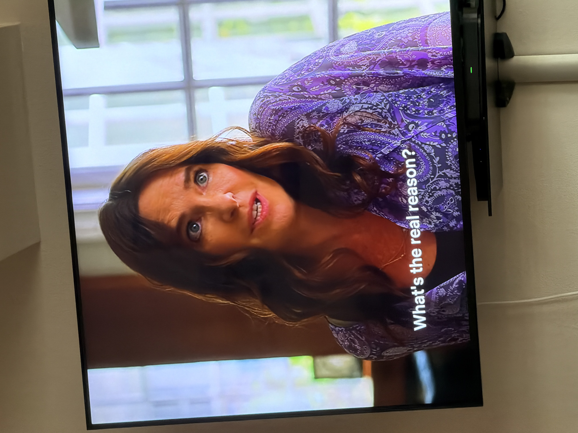 The image is a photograph of a flat-screen television displaying a woman, with the screen rotated approximately 90 degrees clockwise.
The central subject on the TV screen is a woman with long, wavy brown hair, fair skin, and striking blue eyes. Her expression is one of intensity, possibly surprise or concern, with her eyes wide and mouth slightly open as if speaking or reacting. She is wearing a purple garment with an intricate, possibly paisley, pattern. The background displayed on the screen is blurred but shows what appears to be a bright indoor setting with large windows or glass panels.
Overlaid on the screen, in white text, are the words: "What's the real reason?".
Adjacent to the right edge of the television, a black device, possibly a streaming stick or small media player, is visible, distinguished by a small green LED light indicating it is powered on. Below this device, two black wall mounts or cable organizers are attached to a light-colored wall. A white cable also runs horizontally across the wall in the upper right portion of the image. The overall setting is an indoor environment, likely a home, where the television is mounted or positioned against a neutral-toned wall.