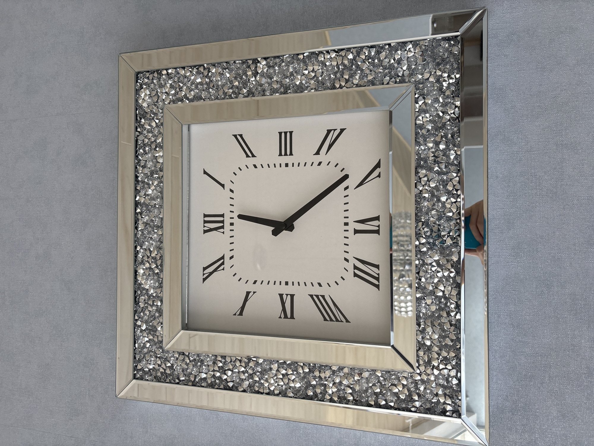 A square, ornate clock with a mirrored frame encrusted with small, clear crystals hangs on a textured gray wall. The clock face is white with black Roman numerals and two black hands. The hour hand points to VI, and the minute hand points to IV. The clock is the primary subject, positioned centrally in the frame, and angled slightly downwards. The background is a plain, light gray wall. There is a reflection in the lower portion of the clock's mirrored frame showing part of a person's arm and hand holding a phone. The image is taken from a slightly elevated angle.