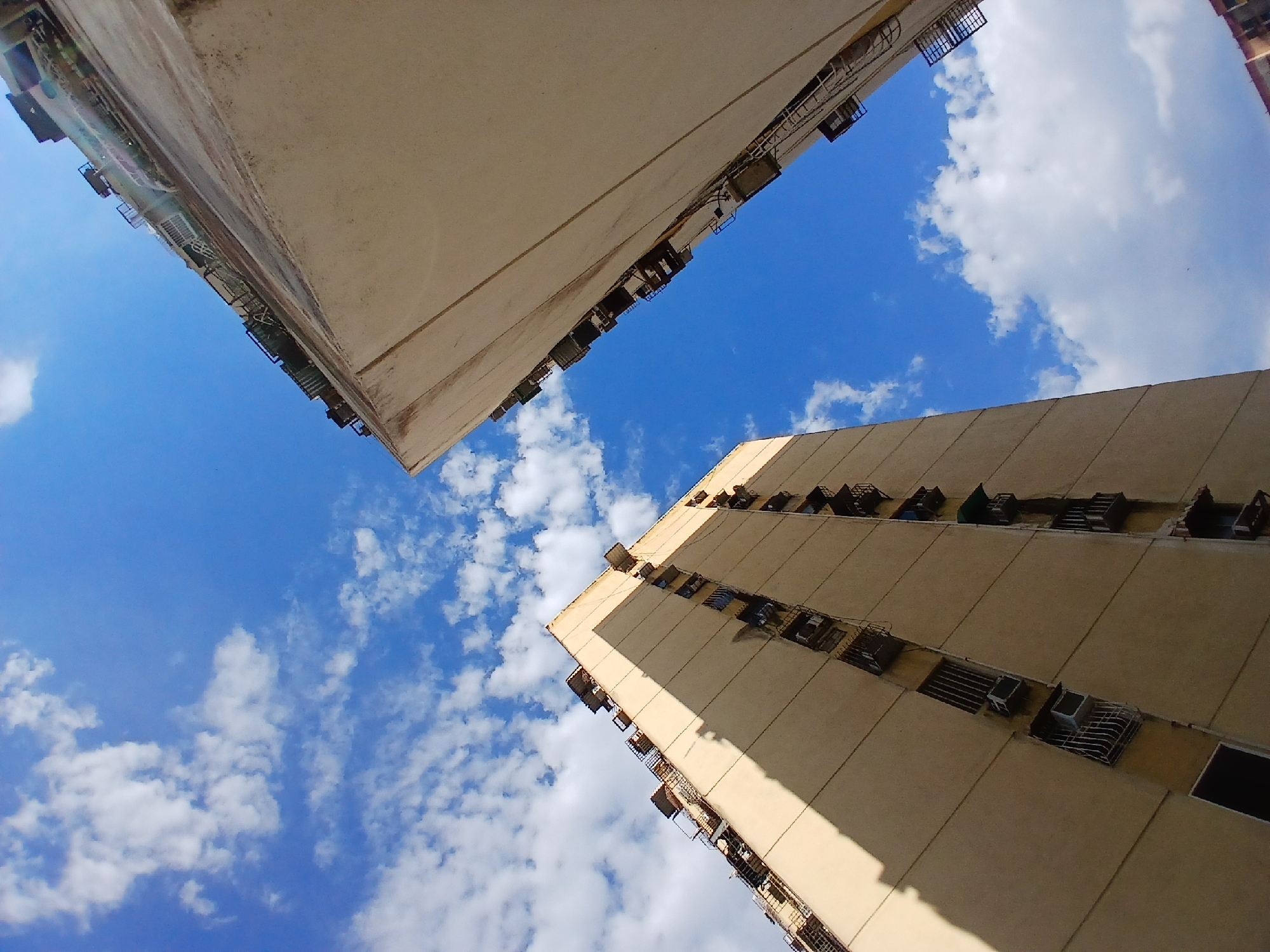 This photograph presents an extreme low-angle, worm's-eye view looking upwards between two tall, multi-story buildings against a bright blue sky. The scene is set in an urban environment, specifically Ciudad Guayana, Venezuela.
The two buildings, positioned on either side of the frame, are light beige or off-white in color, suggesting a concrete or stucco construction. They exhibit numerous small, rectangular openings along their facades, some fitted with metal grates or cages, while others appear to house external air conditioning units or utilitarian structures. The building on the left also shows several balconies with railings. The surfaces of the buildings display some signs of wear or weathering.
The sky dominating the central and upper portions of the image is a clear, vibrant blue, dotted with scattered white, fluffy cumulus clouds. Strong daylight illuminates the scene, casting prominent, distinct shadows on the side of the building to the right, indicating a high sun position, likely midday or early afternoon.
There are no people, visible activities, or discernible text in the image. The focus is purely on the architectural elements and the expansive sky framed between them, highlighting the verticality of the urban landscape.