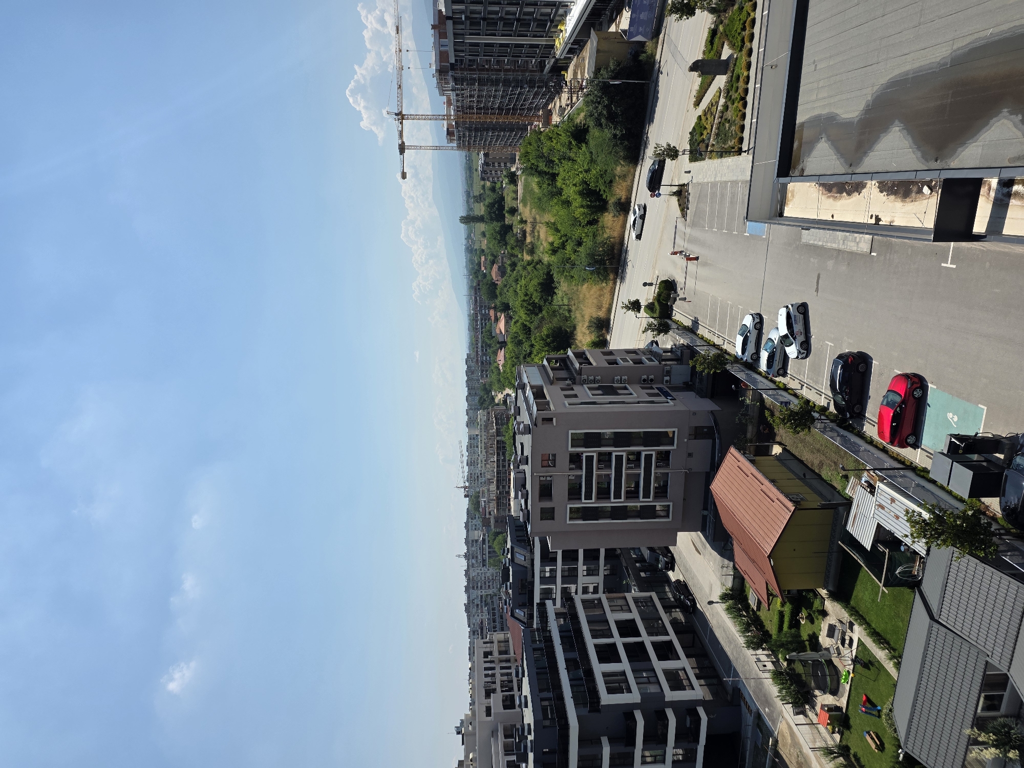 The image captures an aerial view of a modern residential area in Plovdiv, Bulgaria. The scene is dominated by contemporary apartment buildings with dark facades and numerous windows. In the foreground and mid-ground, several multi-story buildings are visible, some appearing to be under construction with cranes present. A street runs through the scene, with a few cars parked in designated spots. To the left, a small park area with playground equipment and a trampoline is situated next to a yellow building with a red roof. The background reveals a sprawling urban landscape with more buildings and a hint of greenery, leading towards distant hills under a clear blue sky with scattered clouds. The environment appears to be a well-developed urban or suburban area.