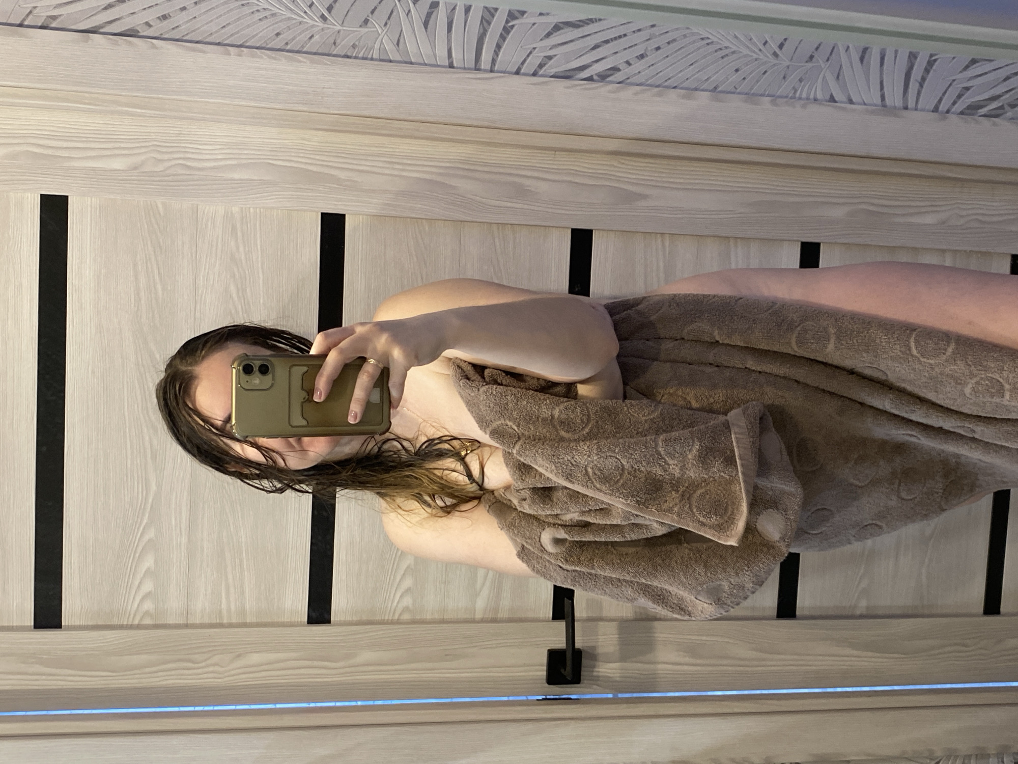 The image depicts an indoor scene located in the city of Noviy, Russia. A person, appearing to be female, is shown from the chest down, with their head partially obscured by a smartphone held in their right hand. The individual's wet, brown hair frames their face, which is behind the phone. They are wrapped in a large, brown, textured towel with circular patterns, covering their torso and extending down their legs. A golden ring is visible on the ring finger of the right hand. A faint necklace with a small pendant is visible on the individual's chest. The background features a light-colored wooden door on the left with two horizontal black accent strips and a black door handle. To the right, a wall is covered in light-toned wallpaper featuring a repeating palm leaf pattern. A vertical strip of light, emitting a blue hue, is visible on the far left edge of the frame. The scene is captured as a selfie, indicating the person is utilizing a reflective surface.