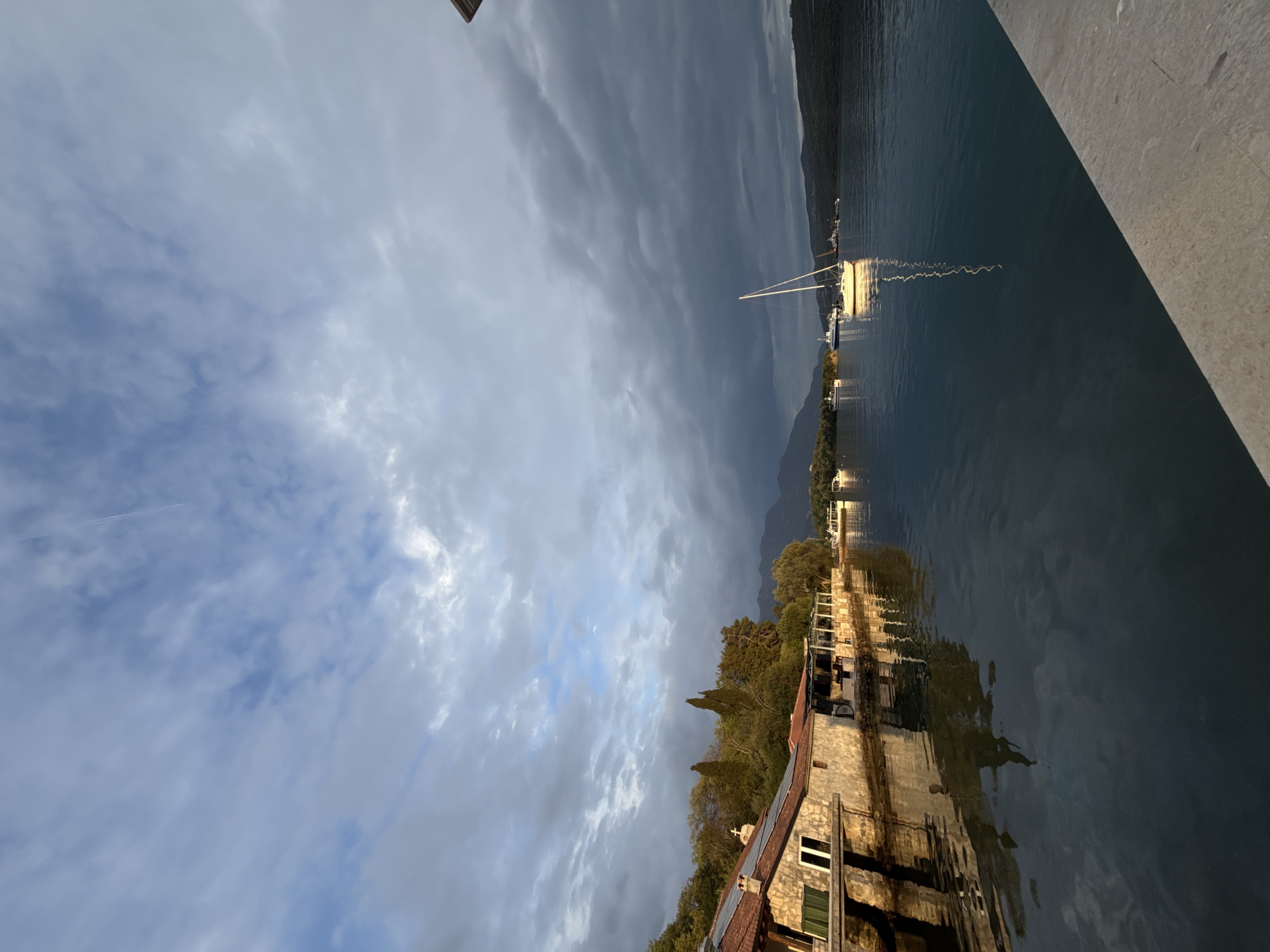 The image shows a landscape in Bogišići, Montenegro. The sky is filled with gray clouds. In the background is a body of water with a sailboat. There are mountains behind the water. The foreground shows a building and some trees. The image is rotated 90 degrees counterclockwise.