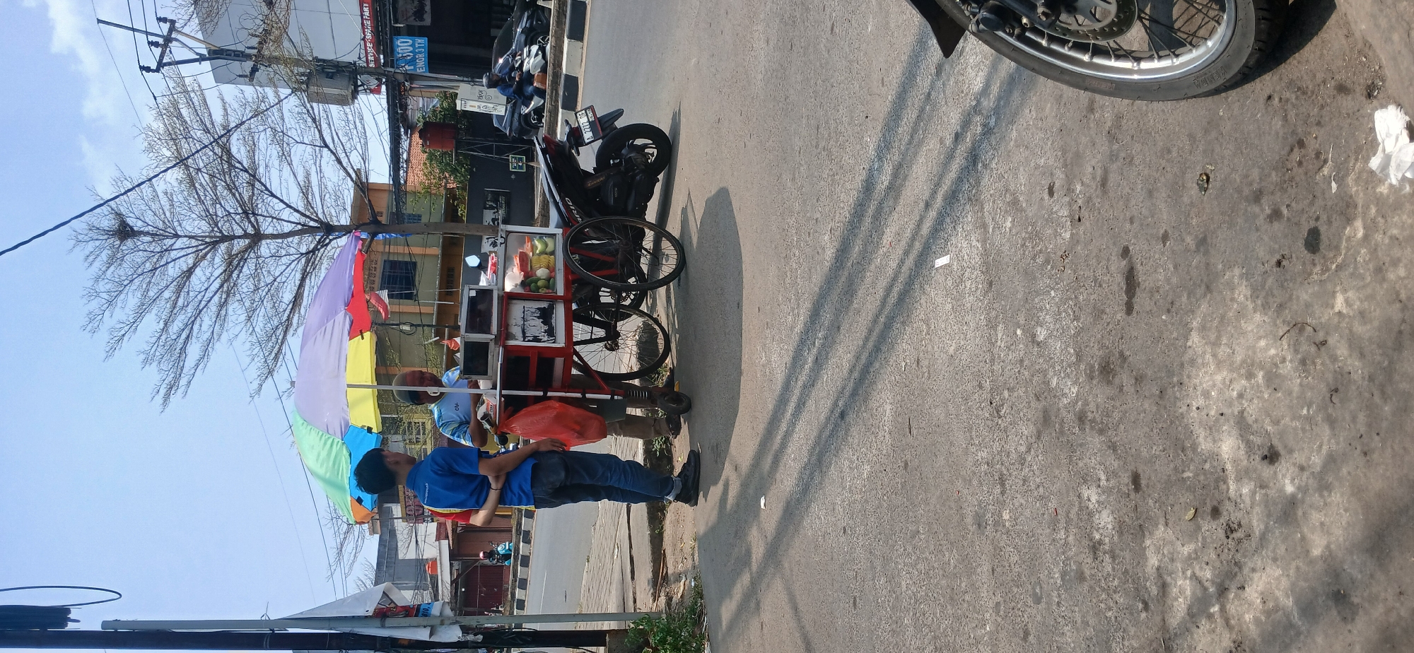 In Baros, Indonesia, on a sunlit day, two individuals stand beside a colorful umbrella-covered food cart. A young man in a blue shirt and jeans stands to the left, his arms behind his back, looking towards the cart. The other individual, an older person in a light blue shirt, is attending to the cart, which displays various food items and appears to be serving customers. A red plastic bag hangs from the cart. Behind them, a motorcycle is parked. The scene unfolds on a paved street next to a sidewalk, with buildings and trees visible in the background, indicating a commercial or residential urban area. Overhead power lines and a utility pole are also present. The overall impression is a typical street scene in Indonesia, likely during daytime business hours.
