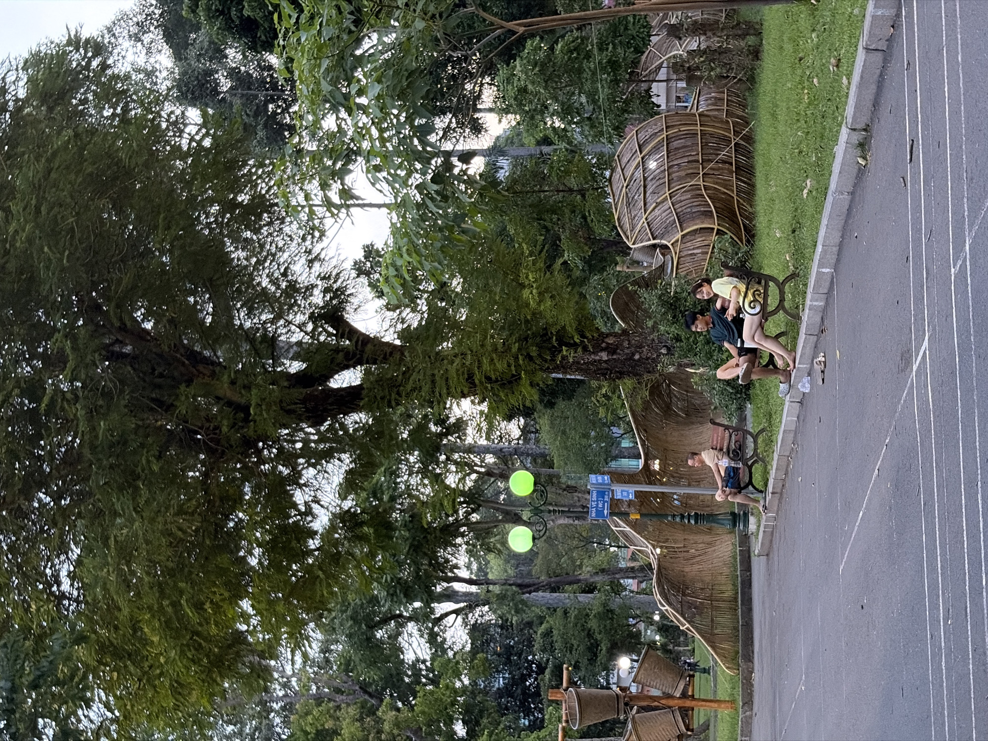 In the city of Thủ Đức, Vietnam, a park scene unfolds with a paved area in the foreground marked with white lines. Two people are seated on a dark-colored park bench to the right of the image. The person on the left is a young man wearing a dark t-shirt and shorts. The person on the right is a young woman wearing a yellow t-shirt with a pattern and dark shorts. They appear to be engaged in conversation, with the woman looking towards the left. Behind them, a grassy area features a unique, curved structure made of woven bamboo-like material, resembling a large creature or abstract sculpture. A lamppost with two bright green lights stands near the bench. Further back, a large tree with dense green foliage dominates the upper portion of the image, with other trees and greenery forming a backdrop. The lighting suggests it is either early morning or late afternoon.