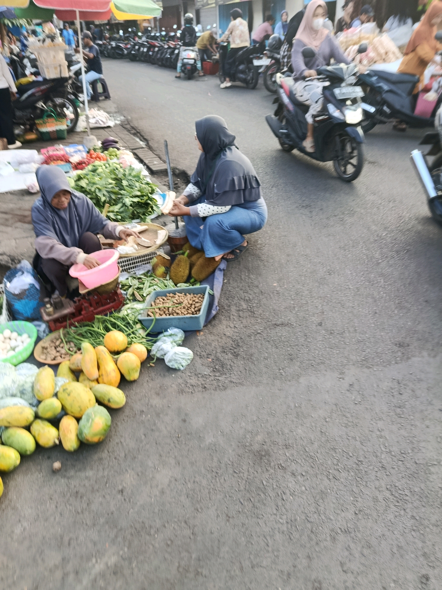 The image depicts a street scene in Bululawang, Indonesia, likely a market area. In the foreground, two women are crouched near a display of fresh produce. The woman on the left wears a gray hijab and a dark jacket over a patterned shirt, with blue pants. She is seated next to a red and silver scale and a pink basin. The woman on the right also wears a hijab, this time a light pink one over a gray top and blue pants. She appears to be interacting with the woman on the left.
A variety of fruits and vegetables are spread out on the ground and in containers. These include papayas, green beans, what appear to be jackfruits, eggs in a green basket, and a blue crate filled with peanuts. In the background, several motorcycles are parked, and people are seen milling about, some on motorcycles. One woman in a pink hijab is riding a black scooter, wearing a face mask. The overall setting suggests a busy roadside market in a town. The ground is paved asphalt, and there are colorful umbrellas providing shade.