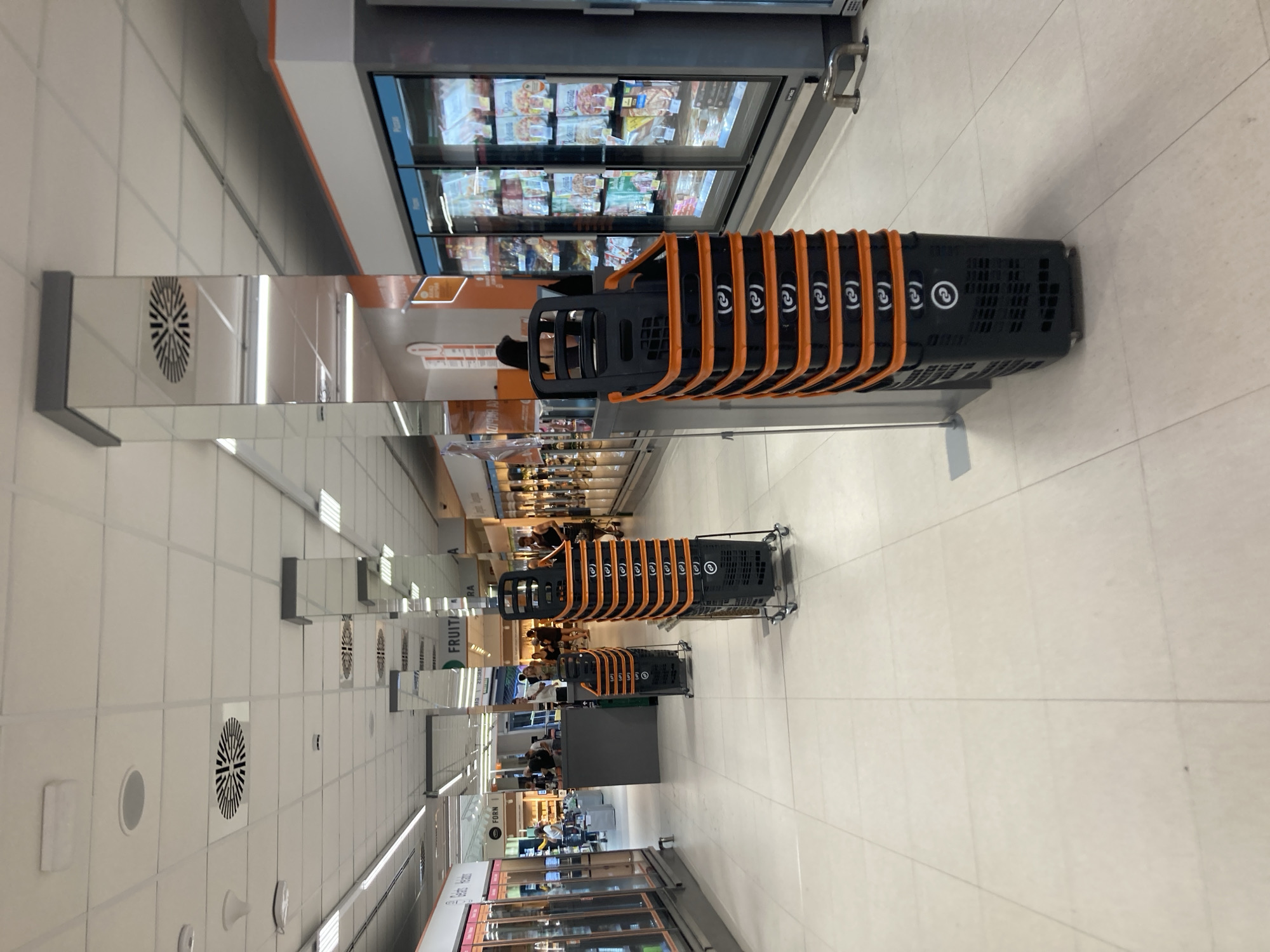 The image depicts the interior of a supermarket aisle, likely in Barcelona, Spain, given the text "FRUITA" (fruit in Catalan). The scene is captured from a slightly elevated perspective, looking down the length of the aisle. The dominant objects are rows of black shopping baskets with orange accents, neatly stacked on trolleys. Several individuals, appearing to be shoppers and possibly staff, are visible in the background, engaged in typical supermarket activities like browsing shelves and pushing trolleys.
Reflective surfaces, possibly overhead signage or polished ceiling elements, create a sense of depth and repetition in the image, reflecting the baskets and the aisle's ceiling lights. The flooring appears to be light-colored tiles. Overhead, ventilation grilles and lighting fixtures are visible.
The time of day is not explicitly indicated, but the artificial lighting suggests it could be any time, though the general atmosphere feels like daytime shopping. There are no strong indicators of weather conditions.
Visible text includes "FRUITA" and "RA" on what appears to be aisle signage, along with "FORN" (oven/bakery in Catalan) further down the aisle. Some of the baskets also have a circular logo with what looks like the number "2" inside, repeated. The overall impression is that of a modern, well-lit supermarket environment. The scene appears calm and ordinary, with no overt emotional expressions discernible from the distant figures.