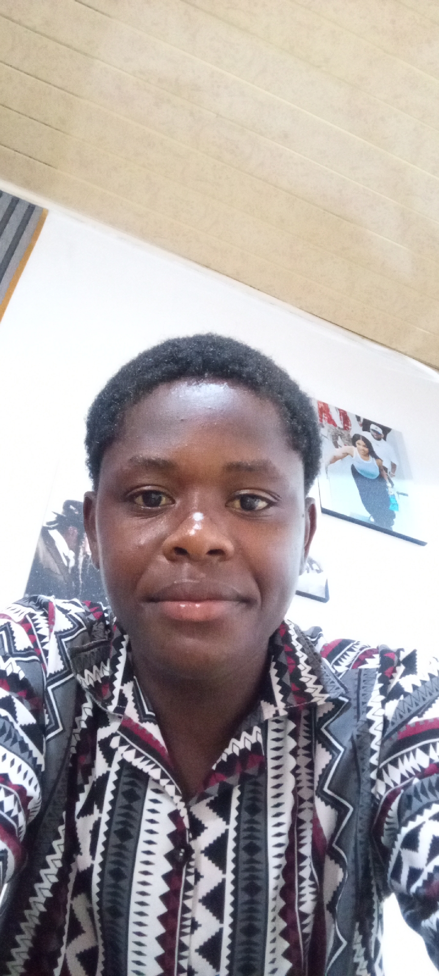 A young man with short, dark hair and dark skin is centered in the frame, wearing a button-down shirt with a geometric pattern of black, white, and maroon. He appears to be indoors. The background features a white wall with two framed images; one is of a woman in a blue dress, and the other is less clear. Above the wall is a beige, paneled ceiling. The location is Unknown, Unknown.