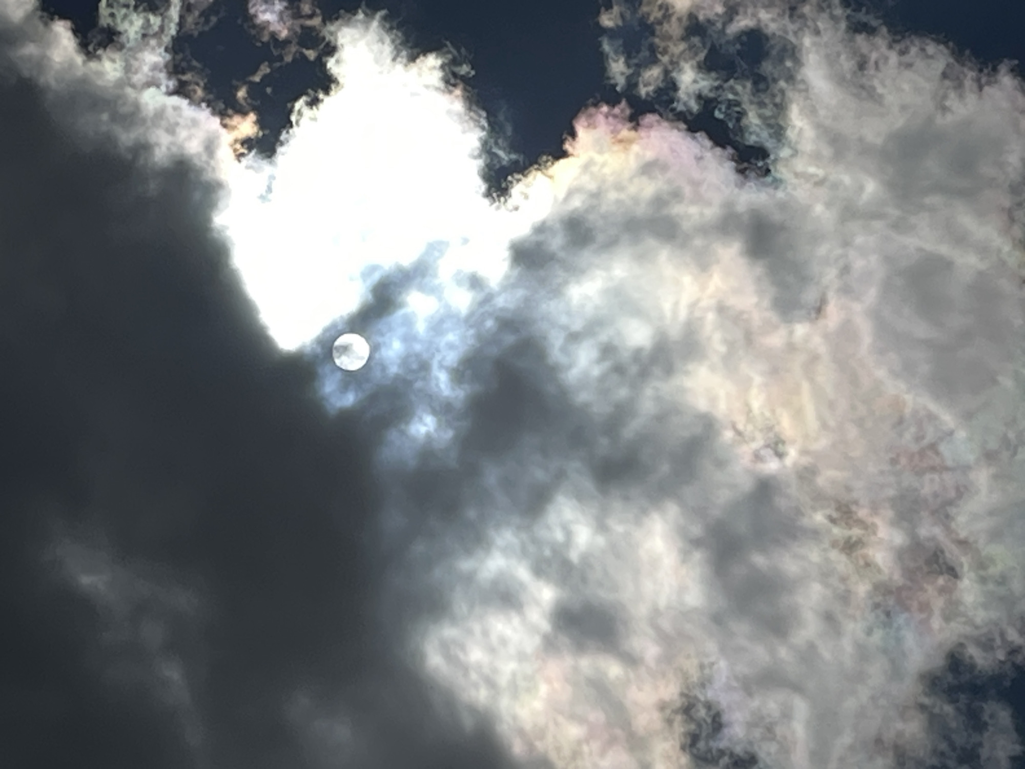 An image captures a sky filled with dramatic clouds, predominantly dark gray and white, with hints of iridescent colors like pink and green appearing where light interacts with the edges of the clouds. A bright, luminous orb, likely the sun, is partially obscured by these clouds, creating a halo-like effect with rays of light emanating outwards. A small, circular, pale disk, possibly the moon, is visible to the left of the sun, appearing faintly white against the darker clouds. The overall impression is of a dynamic and somewhat mystical sky, possibly indicating unusual atmospheric conditions.