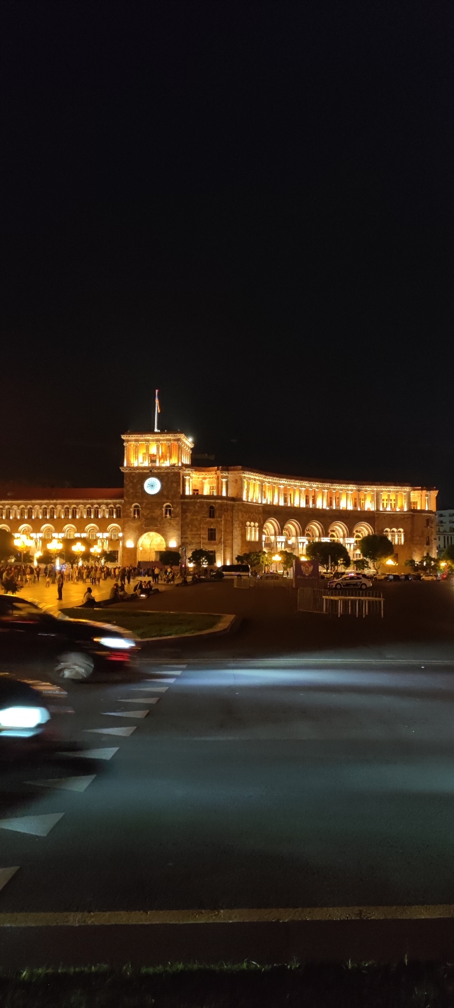 The image depicts Republic Square in Yerevan, Armenia, at night. A large, illuminated building dominates the scene, with a clock tower and a flag atop it. Arches and rows of columns are visible along its facade. Several people are standing or sitting in front of the building. In the foreground, a road with blurred car lights indicates motion. A crosswalk with white triangles is visible on the road. The night sky is dark, providing a backdrop for the illuminated building.