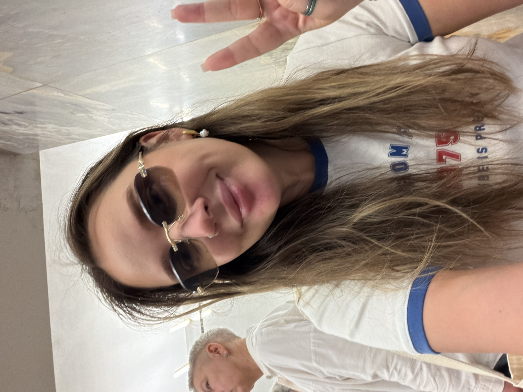 A woman with long brown hair and sunglasses makes a peace sign in what appears to be a selfie. She is wearing a white shirt with blue trim and a t-shirt with "OMBE IS PRE" written on it. A second person is partially visible in the lower left corner of the image. The location is not identifiable from the image.