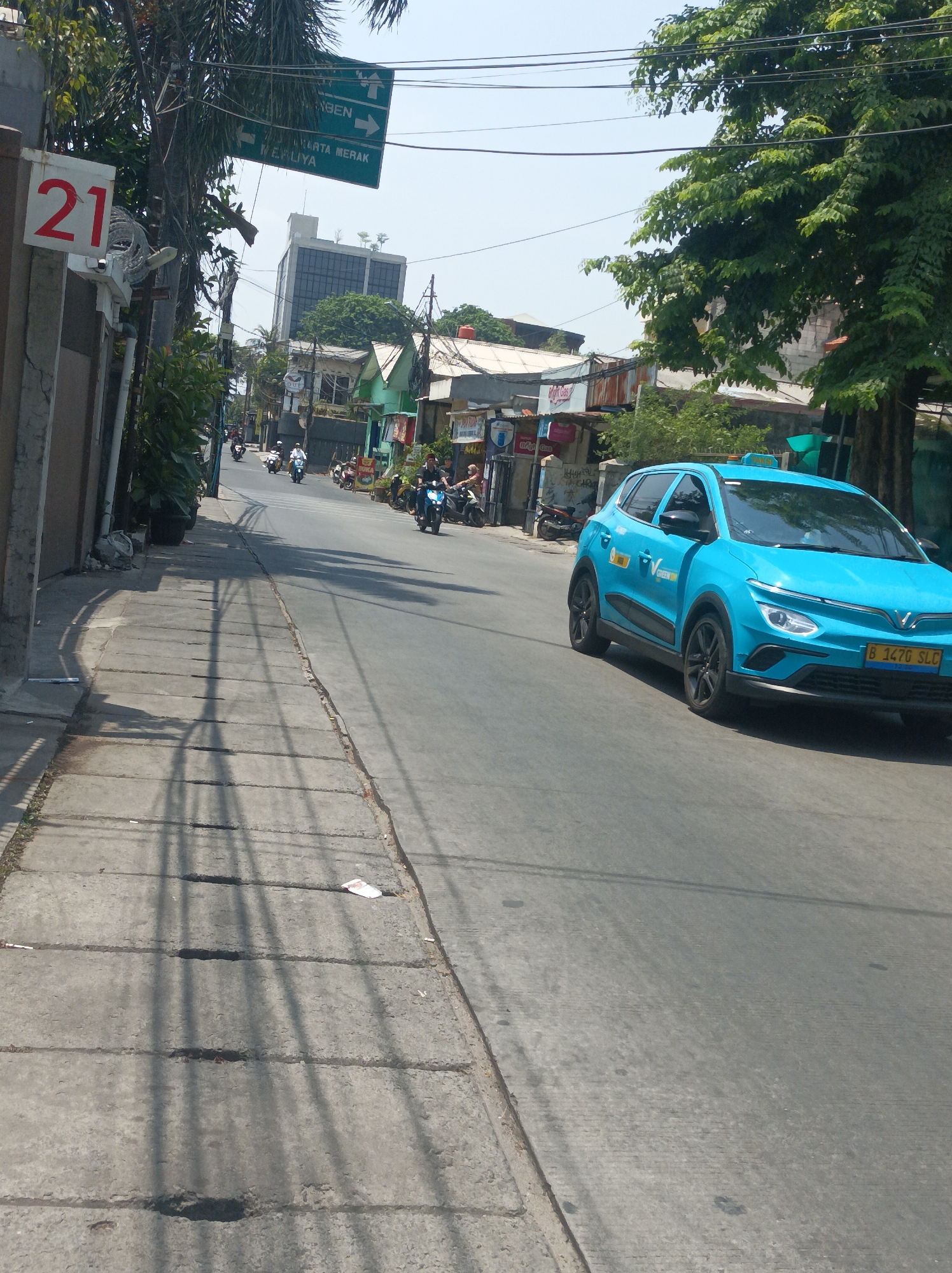 The image captures a street scene in West Jakarta, Indonesia. A bright blue electric car, identified as a taxi with the license plate B 1470 SLC, is driving on the right side of the road. The car has a green logo on its side and a yellow sign on its roof. The road is paved and shows tire marks and some debris. To the left of the road, there is a concrete sidewalk with irregular paving stones and shadows cast by overhead wires and trees. A white sign with the number "21" in red is visible on a pole to the left, near the sidewalk. Further down the street, other vehicles, primarily motorcycles, are visible, with a few people riding them. Along the sides of the street, there are buildings of various sizes, including some low-rise structures with corrugated metal roofs and a taller, modern building in the background. Trees with green foliage are present, and utility wires crisscross the sky. Traffic signs are also visible, including green directional signs pointing to "TANGERANG" and "JAKARTA MERAK". The sky is clear and bright, suggesting a sunny day.
