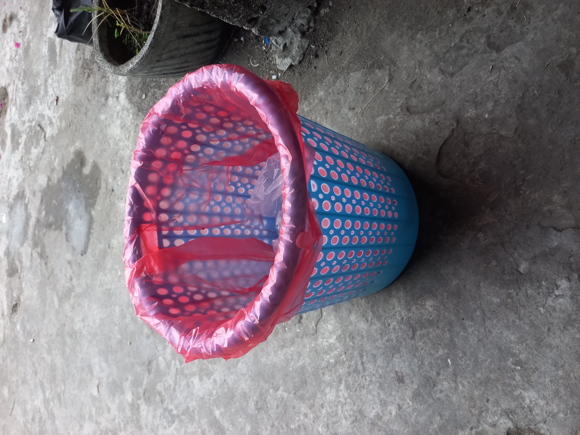 An outdoor scene, likely in Kebon Agung, Indonesia, features a blue plastic trash bin with a red plastic liner. The bin has a perforated design with pink circles. It is situated on a textured concrete surface, possibly a pathway or patio. In the background, a gray concrete planter with a small green plant and dark soil is visible, along with some debris and a black plastic bag. The lighting suggests it is daytime.
