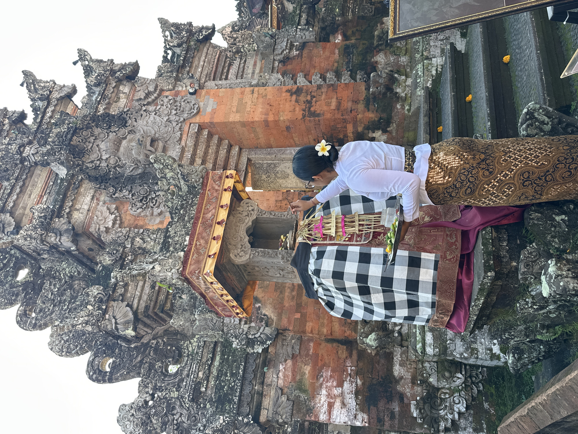 A woman in traditional Balinese attire, a white long-sleeved shirt and a gold and brown patterned sarong, stands at the entrance of a stone temple. She wears a white flower in her hair and glasses. The woman is holding a silver tray with offerings and is in the process of placing something on a statue or altar in front of the temple. The statue is draped with a black and white checkered cloth and a dark red fabric. The temple is constructed of red brick and intricately carved grey stone, with ornate architectural features. Stone steps lead up to the temple entrance, with small yellow flowers scattered on them. The environment appears to be outdoors with an overcast sky. The scene is located in the city of Ubud District, Indonesia.