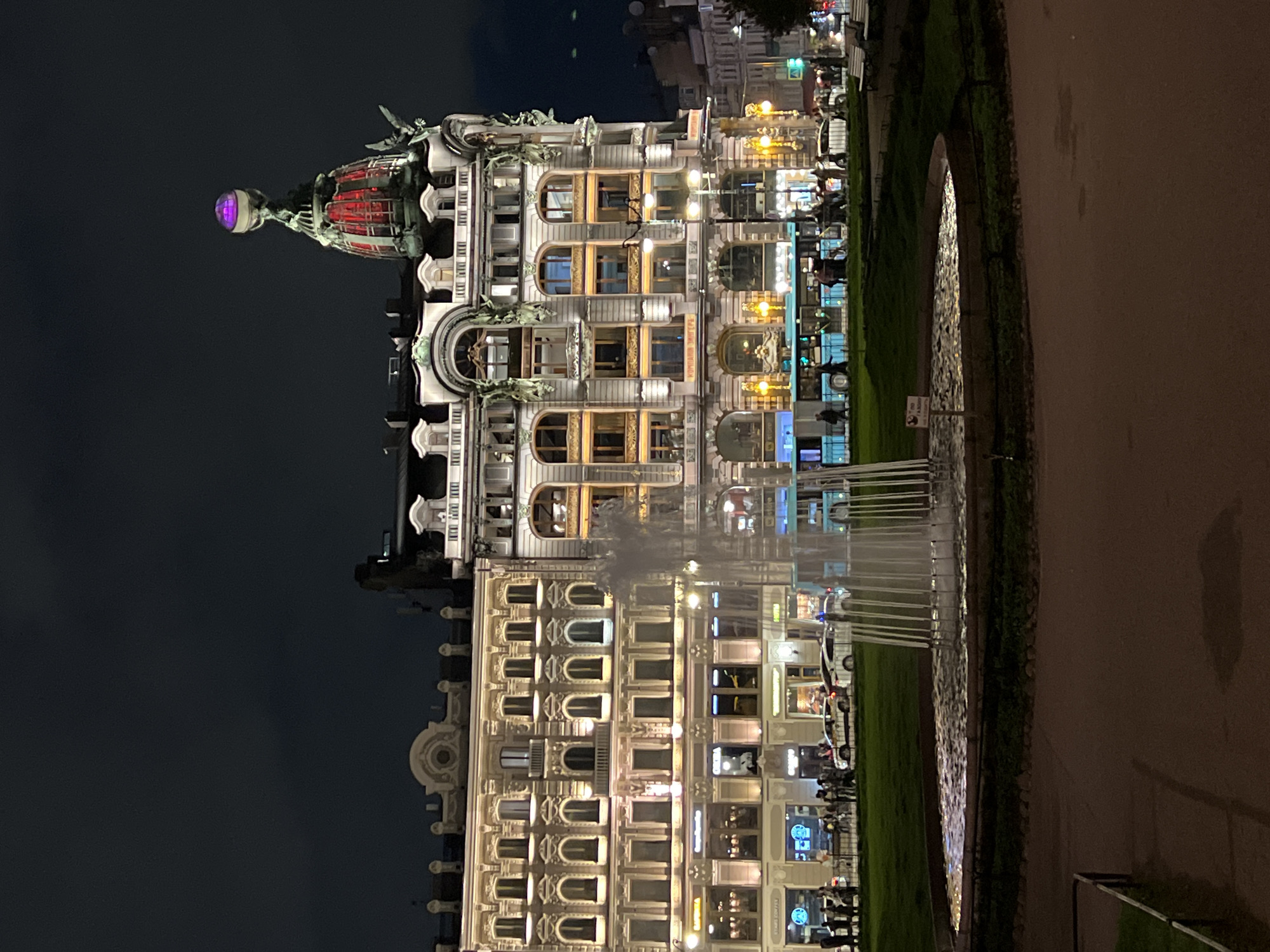 At night, the Singer House (Dom Knigi) stands tall on Nevsky Prospekt in Saint Petersburg, Russia, illuminated by numerous lights. The building's ornate facade features multiple windows with illuminated interiors. A dome topped with a glass globe and statues sits on one corner. Below, a park with benches is visible. A fountain is in front of the building. Pedestrians are walking along the street. The sky is dark.
