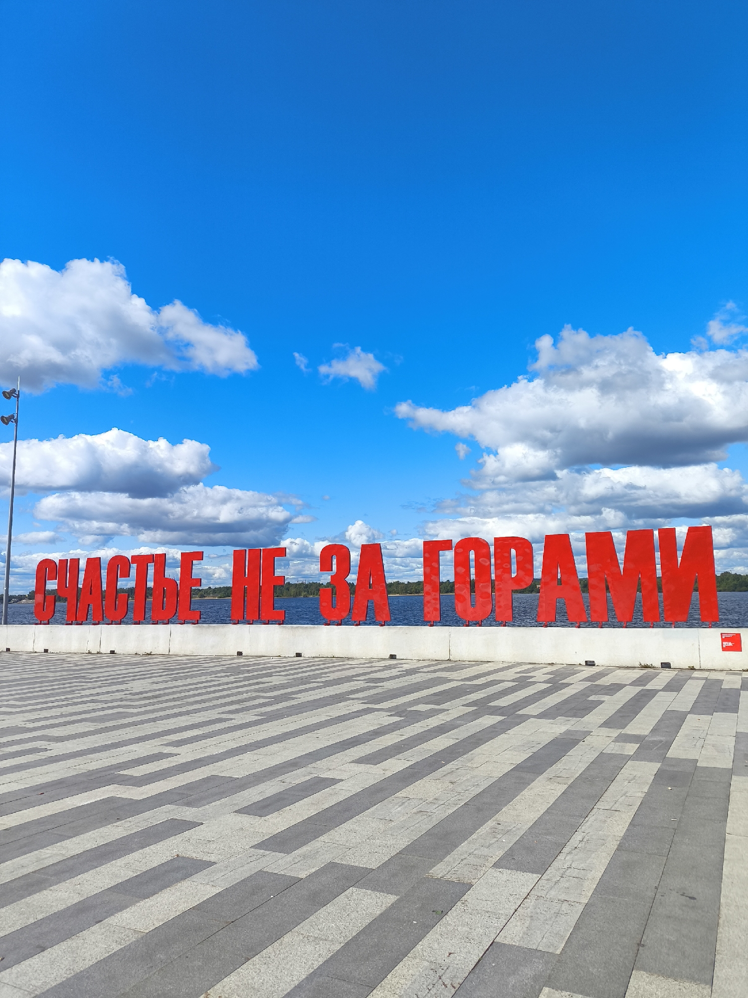 The image shows a large, red, three-dimensional Cyrillic inscription that reads "СЧАСТЬЕ НЕ ЗА ГОРАМИ" (Happiness is not far away). The inscription is situated along a waterfront promenade in the city of Perm, Russia. The promenade has a patterned pavement made of grey and light-colored rectangular tiles. Behind the inscription, a body of water, likely the Kama River, is visible, with a treeline on the far bank. The sky above is bright blue with scattered white cumulus clouds. A lamppost with multiple light fixtures is visible on the far left of the image. There are no people or animals in the frame. The overall scene is a public outdoor space on a sunny day.