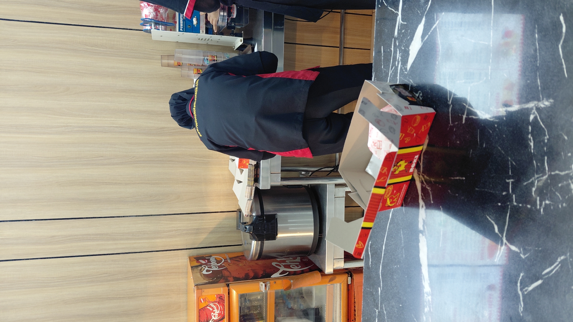 A person wearing a black and red uniform and a head covering stands behind a stainless steel counter in what appears to be a fast-food establishment in Rejasari, Indonesia. The person is facing away from the camera, with their hands near a beverage dispenser. In the foreground, a partially opened red and yellow fast-food box rests on a black marbled surface. Behind the counter, a large stainless steel rice cooker is visible, along with stacked cardboard boxes and various drink containers. The background features a light-colored wood-paneled wall.