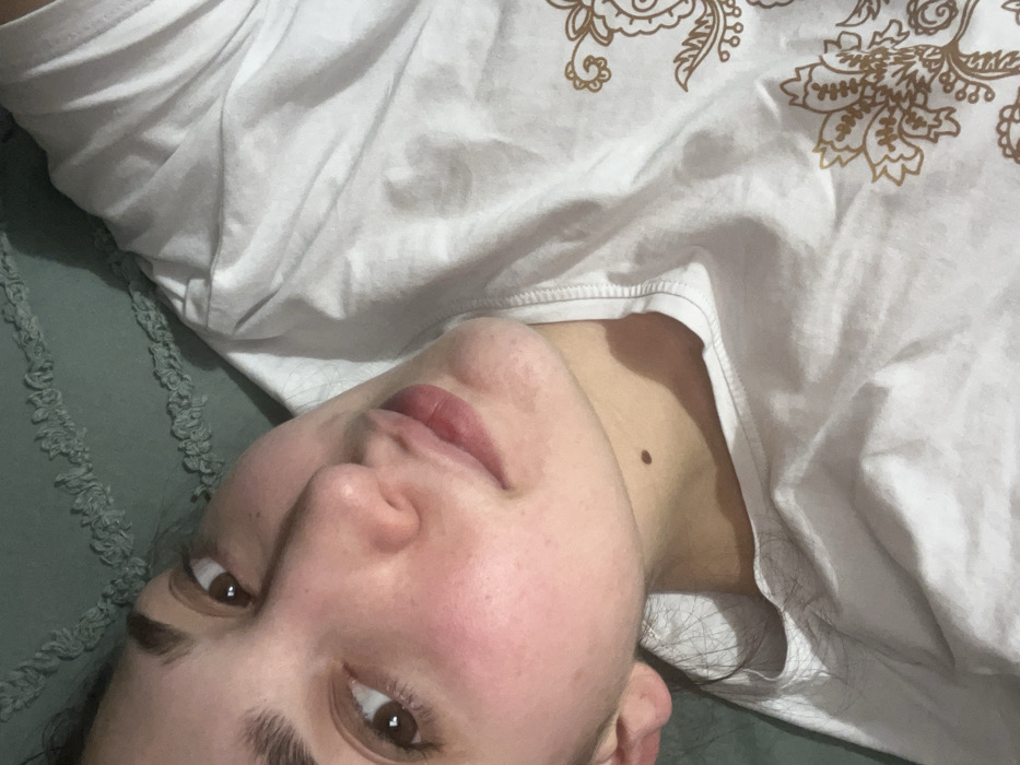 The image shows a person with light skin lying on a green textured surface. The individual wears a white t-shirt with an ornate gold-colored pattern on the upper portion. The person's face is angled towards the camera, revealing brown eyes, dark eyebrows, a small mole on the neck, and pinkish lips. The person appears relaxed, possibly resting or posing for the photograph. The lighting is soft, casting subtle shadows across the face and t-shirt.