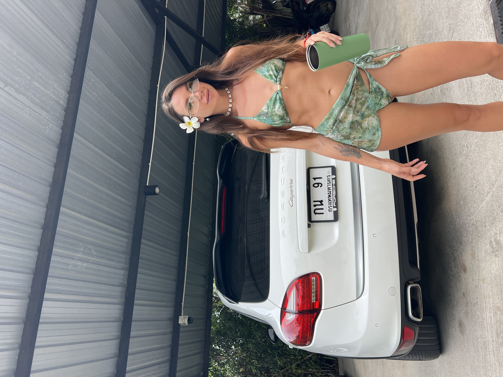 A woman stands in front of a white Porsche Cayenne. The car is parked under a metal awning with visible support beams and a security camera. The woman is wearing a green bikini and a sheer green sarong. She has a white flower tucked behind her ear and is holding a green tumbler. A motorcycle is visible in the background to the right. The license plate on the car appears to be in Thai. The scene is outdoors, with trees and foliage visible behind the car. The ground is paved with concrete.