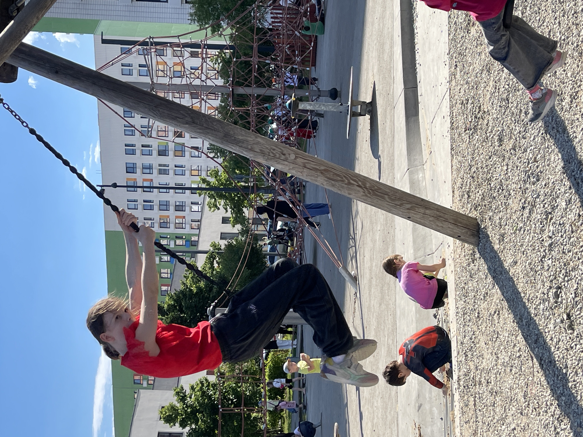 The image captures a vibrant outdoor playground scene in Kommunarka, Russia, under a clear, sunny sky with a few wispy clouds. The bright daylight casts distinct shadows, notably a long shadow from a wooden beam stretching across the light gravel surface.
The primary subject is a child, wearing a red t-shirt and dark pants, actively swinging or using a zip line. They grip a black handle attached to a sturdy chain descending from a thick wooden support beam. Their light brown hair is swept back, and their expression is focused on the activity.
Several other children are visible in the playground. In the mid-ground, a child with long brown hair and a pink top is seen crouching or playing in the gravel. Closer to the foreground, another child in a red and blue top (resembling a Spider-Man suit) and dark pants is also crouching in the gravel. The legs of another child in gray pants and sneakers with pink accents are visible in the upper right. Further in the background, multiple children are engaged on a large climbing net structure made of red ropes and metal poles, with some colorful panels integrated into its design.
The playground features a mix of surfaces: paved concrete areas and a large expanse of light-colored gravel. In the background, modern residential buildings with multiple windows, some featuring colorful frames (orange, green, blue), rise against the sky. One building has a distinctive green upper section. Green trees are scattered around the playground area, adding natural elements to the urban setting.
No visible text is discernible in the image. The overall atmosphere is one of lively outdoor play.