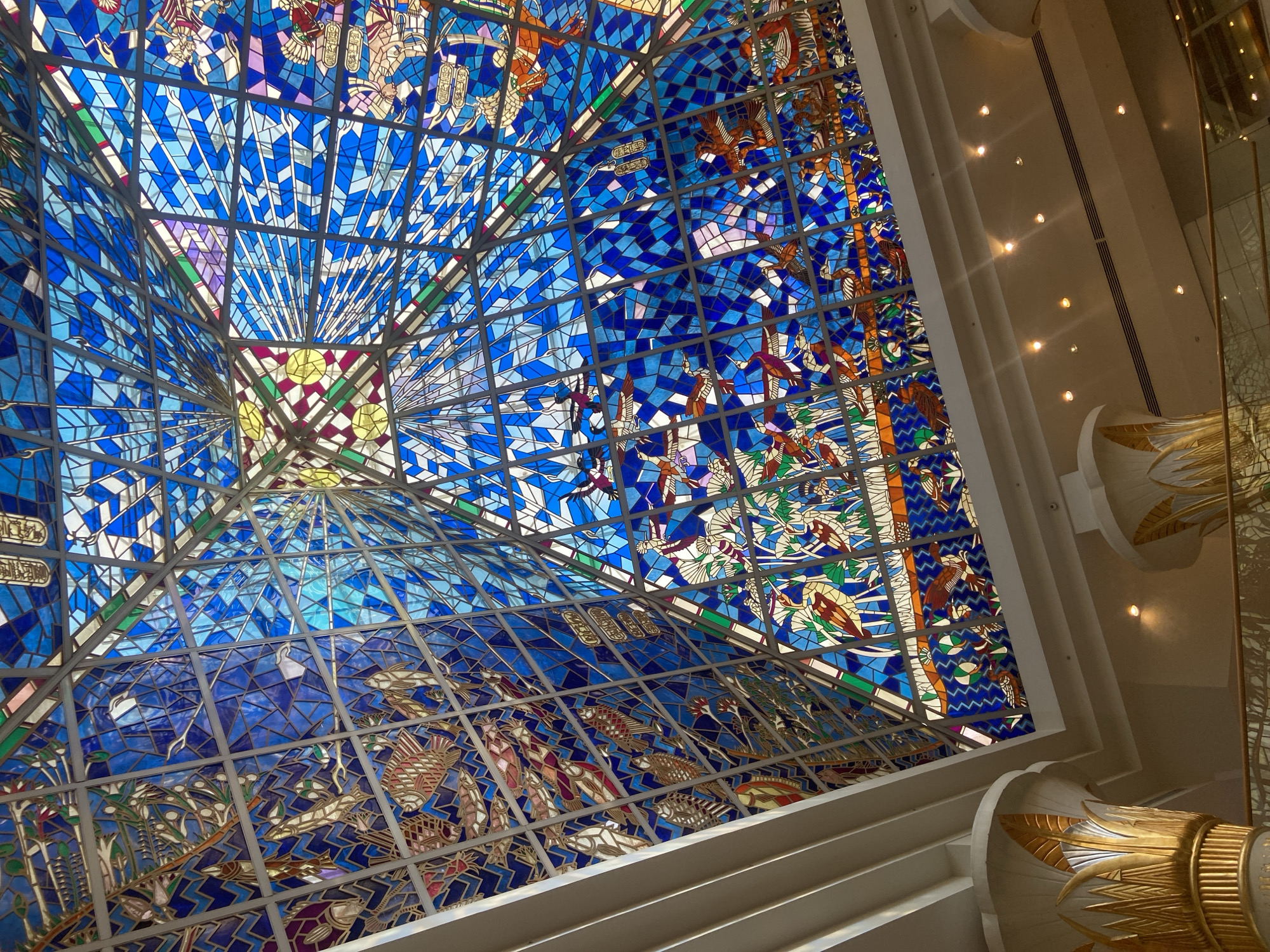 The image shows a stunning stained glass ceiling designed in a pyramid shape, reminiscent of ancient Egyptian architecture. The ceiling is adorned with intricate depictions of various creatures, including birds and fish, set against a backdrop of vibrant blue and geometric patterns. The stained glass is supported by a metal framework, creating a mosaic effect.
Below the stained glass ceiling, the scene reveals the interior of a grand building. Two large, ornate pillars, decorated with gold leaf in a lotus flower design, are visible on either side of the frame, suggesting an opulent and possibly historical or themed environment. The pillars have a fluted design and are topped with decorative elements that resemble Egyptian papyrus. The lighting within the space is warm and diffused, with recessed ceiling lights visible. The overall impression is one of grandeur and artistry, with a clear Egyptian influence in its design elements.
This detailed description strongly suggests the location is the Wafi Mall in Dubai, United Arab Emirates. Wafi Mall is renowned for its architecture, which is heavily inspired by ancient Egyptian design, including pyramid-shaped structures and decorative elements like lotus flowers. The stained glass ceiling, featuring intricate artwork and a pyramid form, is a signature element of the mall's interior design.