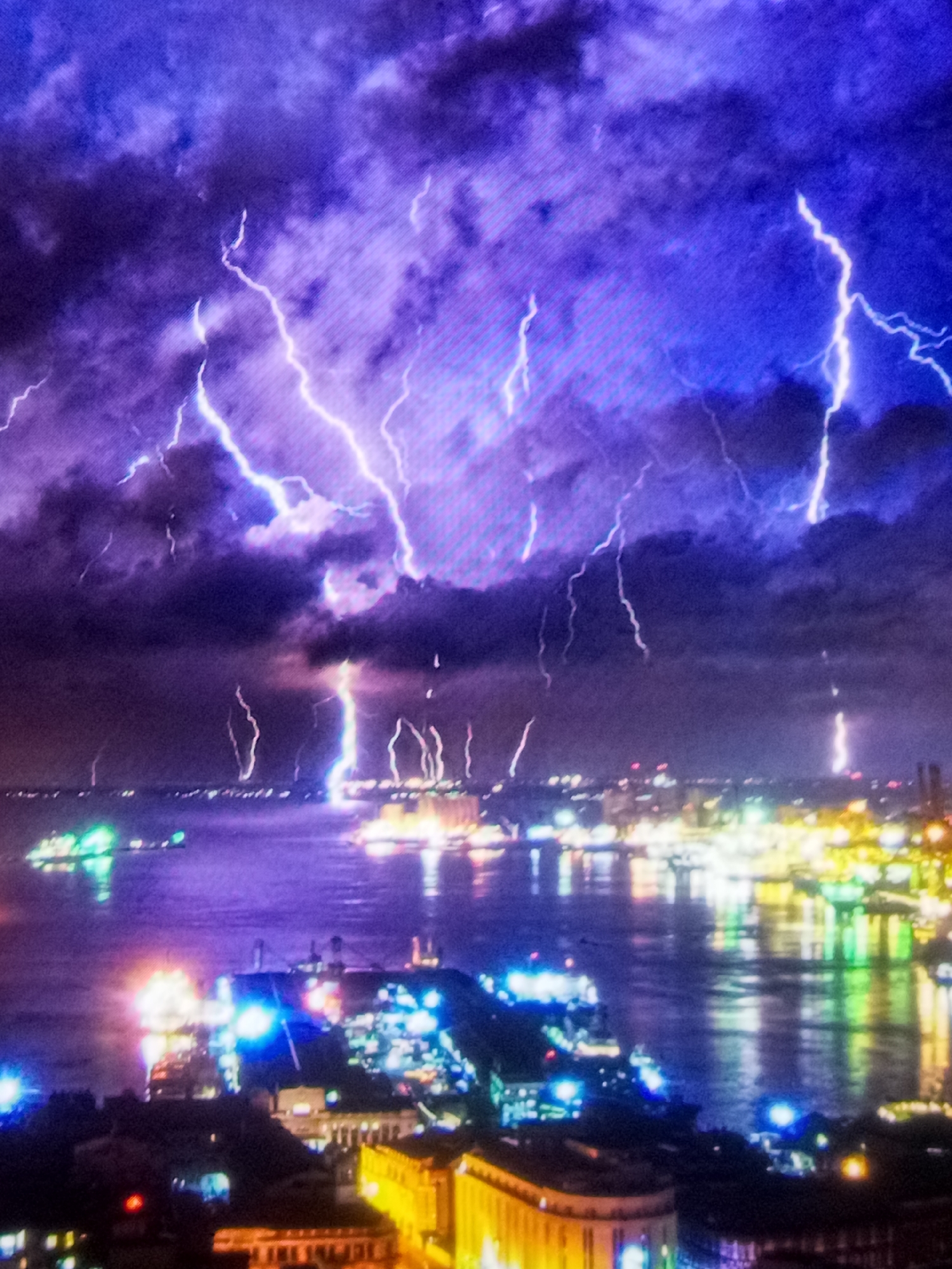 This image captures a dramatic nighttime scene of a city skyline during a powerful thunderstorm. The sky is filled with a vibrant display of lightning bolts, illuminating the dark, swirling clouds with brilliant white and electric blue light. The lightning strikes are concentrated in the upper portion of the image, creating a sense of intense energy.
Below the stormy sky, a city landscape is visible. The lights of the city twinkle and reflect on the calm water in the foreground, creating a blurred yet vibrant panorama. Buildings and structures are silhouetted against the illuminated sky, and the reflections on the water suggest a harbor or bay area. The overall impression is one of a city experiencing a fierce but awe-inspiring weather event. The image's style is somewhat abstract due to the visual effects of the lightning and the nighttime setting.