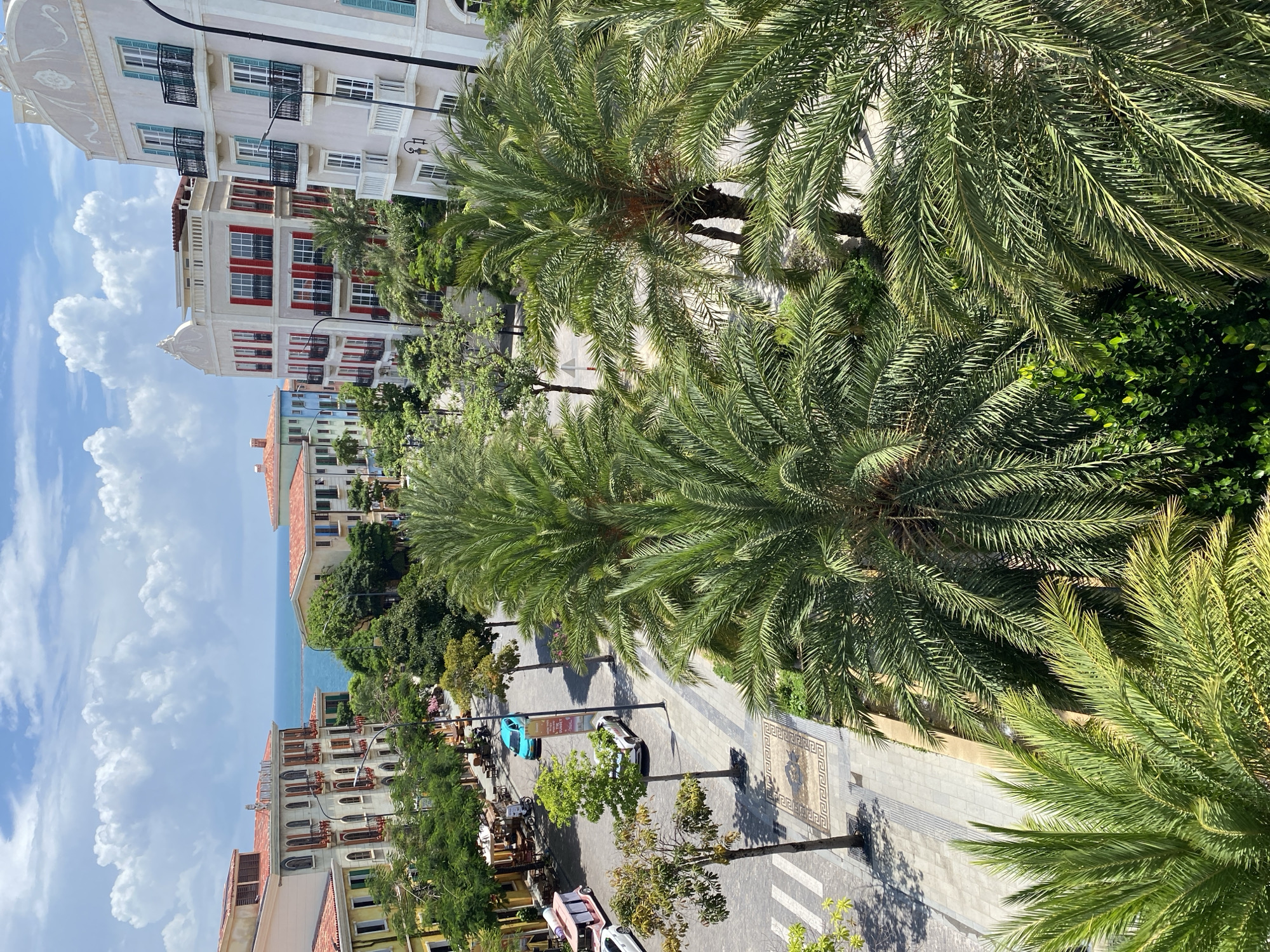 This is a high-angle, full shot of a street scene in the city of Phú Quốc, Vietnam. The scene is dominated by lush palm trees in the foreground, their fronds casting shadows on a paved street and sidewalk. Several multi-story buildings with Mediterranean-style architecture line the street, featuring red-tiled roofs, balconies with black railings, and brightly colored facades. The buildings are a mix of pastel yellow, pink, and white, with some featuring striking red shutters.
On the street, a bright turquoise car is parked, and a white car is visible further down. There are streetlights and lampposts along the sidewalk. Some trees are planted along the street, interspersed with the palm trees. A few outdoor seating areas with tables and chairs are set up in front of some of the buildings. In the background, the blue sea is visible under a bright sky with fluffy white clouds. The overall impression is a vibrant, resort-like atmosphere.