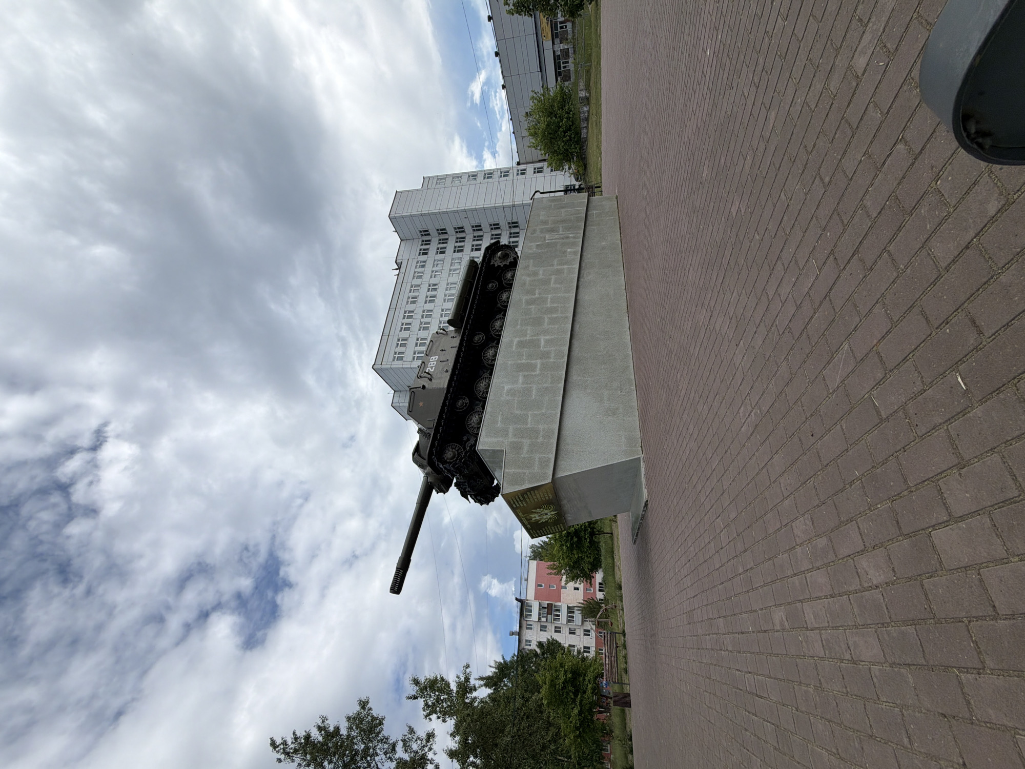 The photograph captures a war memorial in Chaikovsky, Russia, featuring a Soviet-era tank prominently displayed on a concrete pedestal. The tank, painted dark green, has the number "288" visible on its side. The pedestal is angular and gray, with a plaque on its front bearing an emblem and text, likely a dedication.
The memorial is situated in an open, paved area with brick-like stones. In the background, a multi-story white building with numerous windows stands out, suggesting an urban setting. To the left of the memorial, trees and a residential building with a pink and white facade are visible. Further in the distance, other buildings and trees can be seen. The sky is filled with dramatic clouds, with patches of blue sky peeking through. The lighting suggests it is daytime, possibly overcast. There are no people clearly visible in the image, although the presence of a bench and trash can hints at public use of the space. The perspective is low-angle, emphasizing the monument's scale against the sky and buildings.