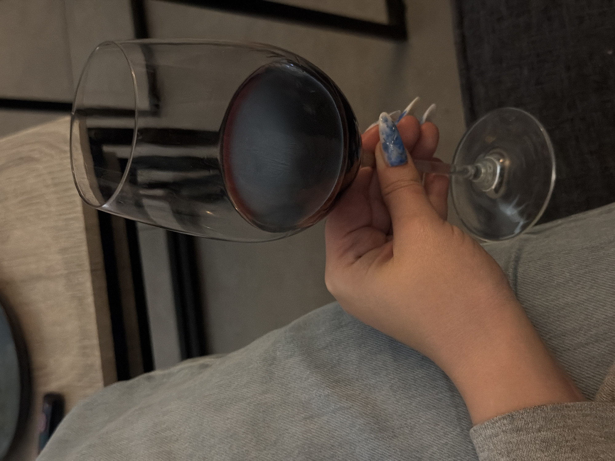 A hand with long, pointed nails with blue-and-white marble polish holds a wine glass filled with red wine. The person is wearing light blue denim pants and a grey long-sleeved shirt. The background shows a grey surface, a black geometric design, and what appears to be part of a table setting with a plate. It is not possible to determine the location in Ramat Gan, Israel based on this image.