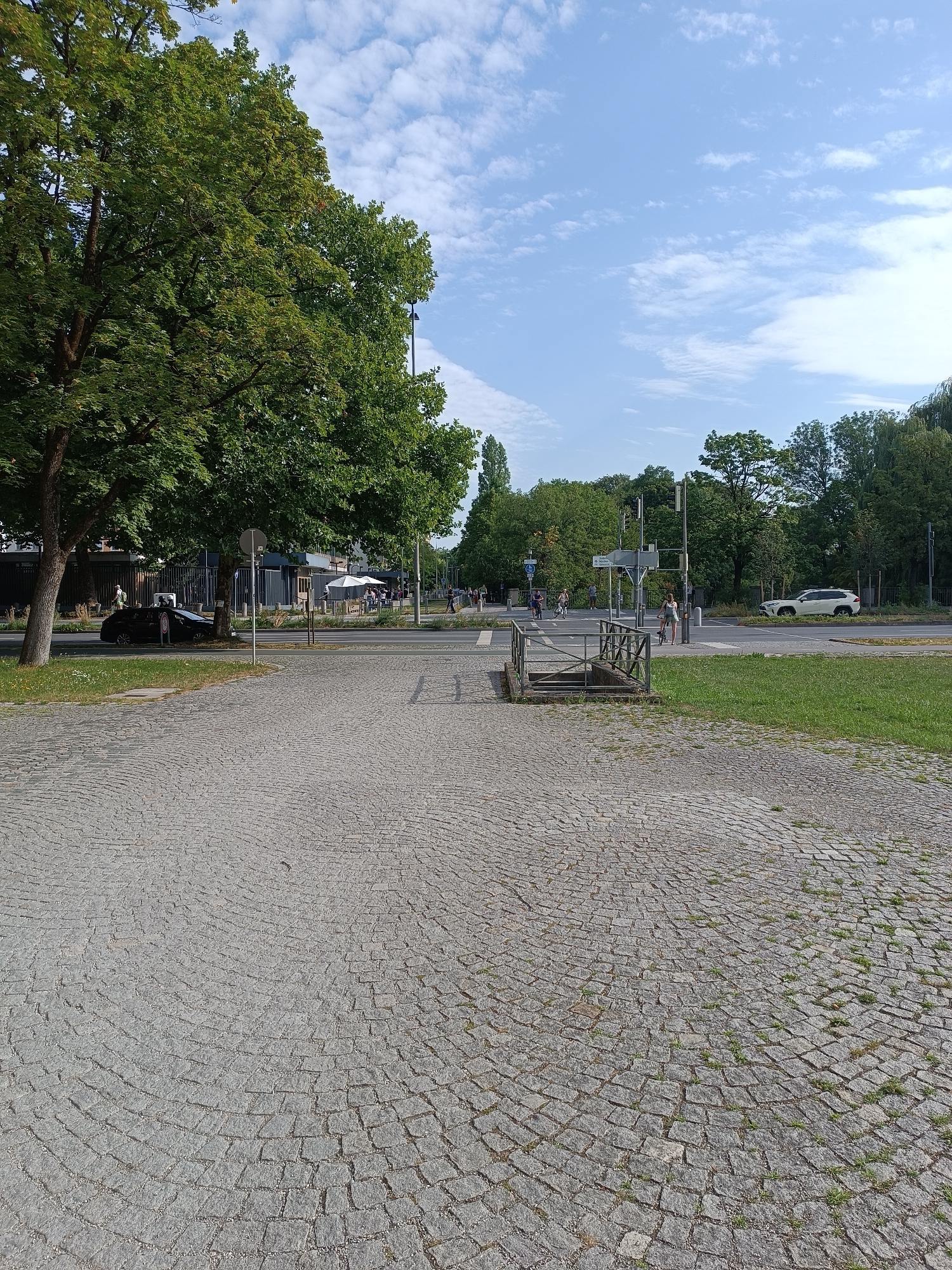 This is a medium-angle shot of a street scene in Munich, Germany, on a clear day with scattered clouds. The foreground is dominated by a cobblestone plaza that curves towards the right side of the frame. A large, leafy green tree stands on the left side of the image, casting some shade onto the cobblestones. Beyond the plaza, a street is visible with traffic, including a black car parked near the left edge and a white SUV further down the road on the right. There are people walking and cycling on the sidewalk and crossing the street. A set of stairs leading down into an underground passage is visible in the middle ground, framed by a metal railing. Buildings and more trees form the background. The overall impression is of an urban environment with green spaces and pedestrian activity.